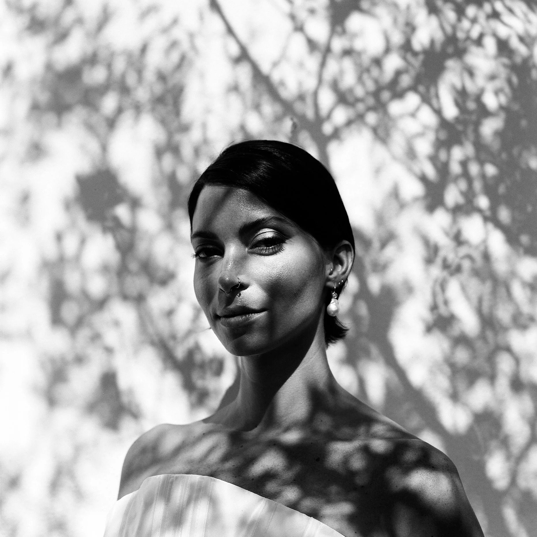 Ritratto di donna con capelli corti e trucco, con ombre di foglie sul volto e sullo sfondo in bianco e nero.