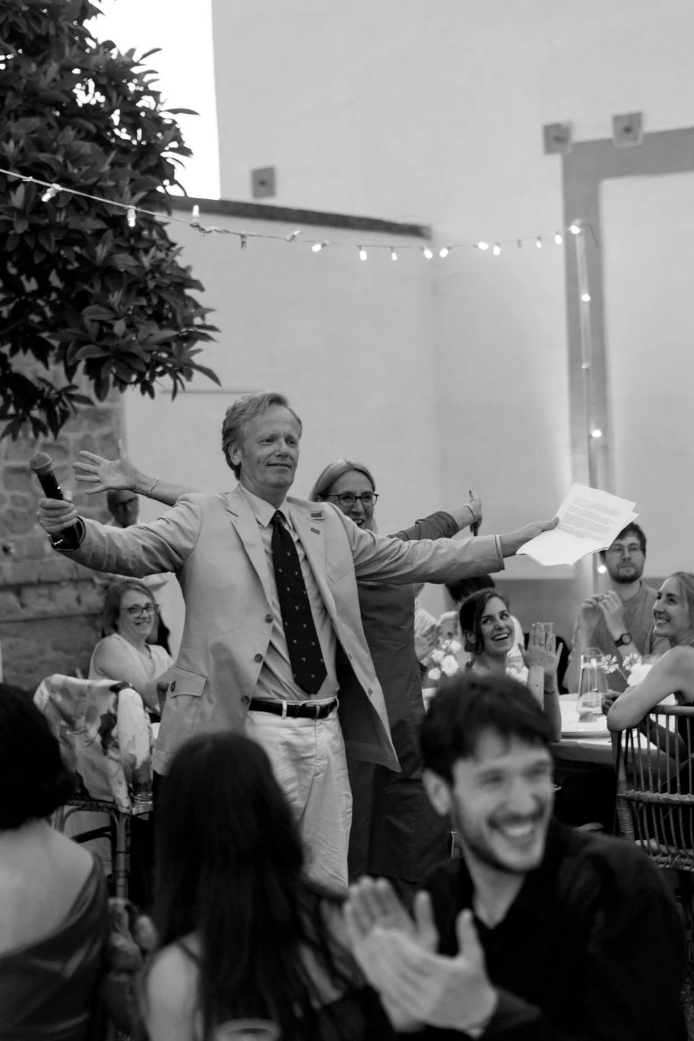 Persona che tiene un microfono e un foglio di carta, con amici in un ambiente festoso all'aperto, decorato con luci