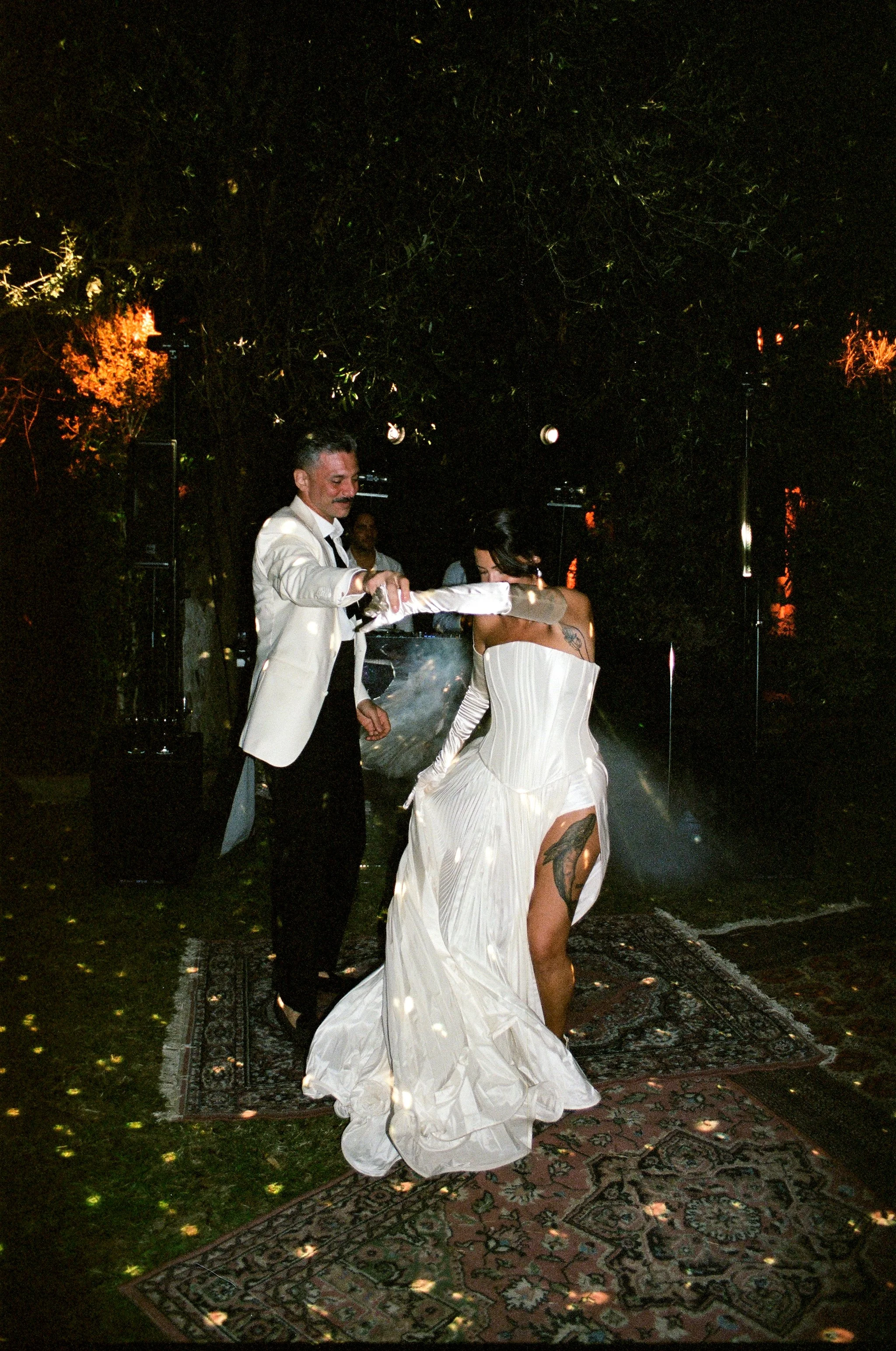 Una coppia di sposi danza durante il loro matrimonio, l'uomo indossa uno smoking bianco e la donna un matrimonio lungo bianco con una gonna ampia e una spalla scoperta, sul pavimento ci sono due tappeti.