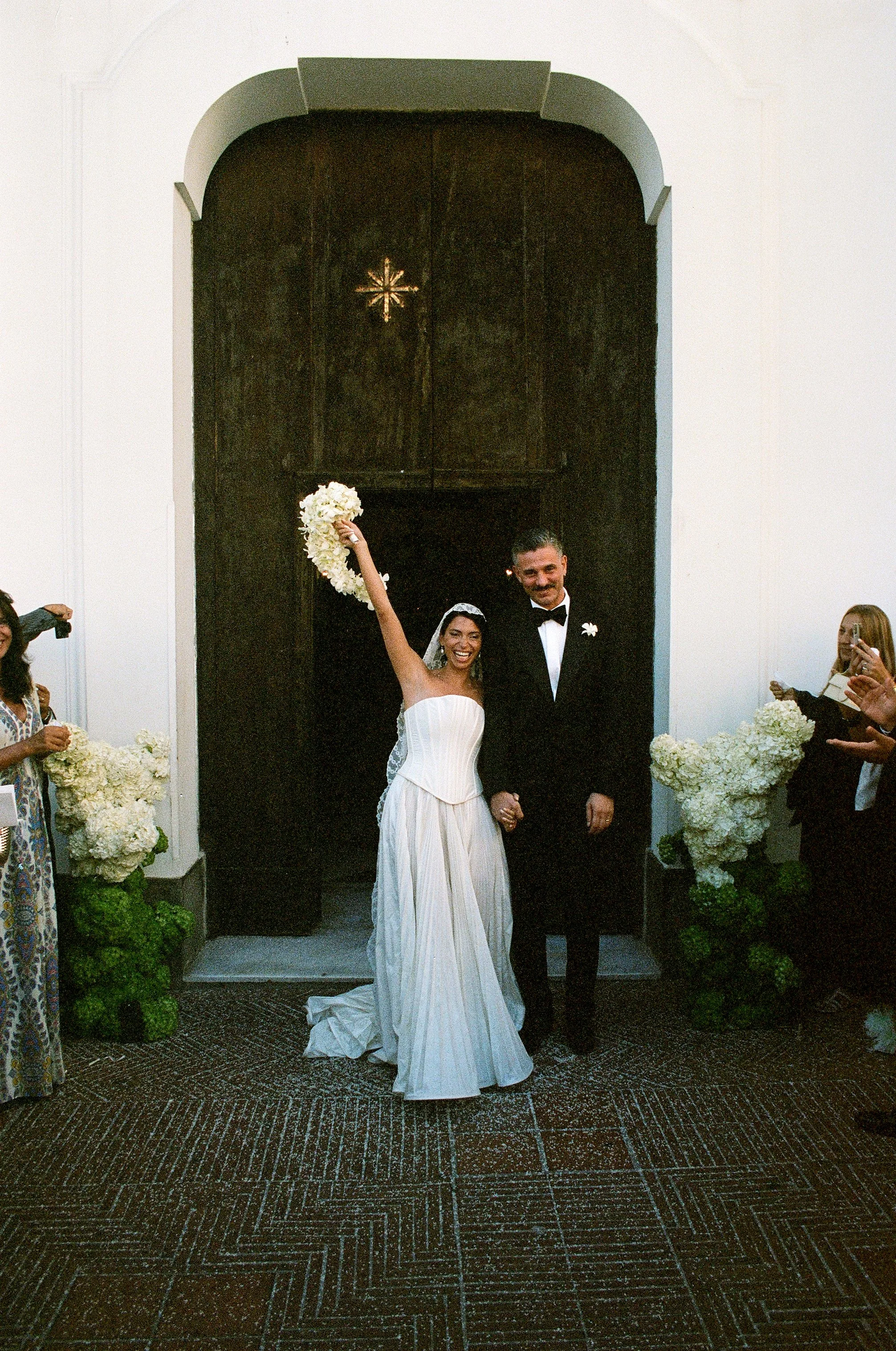 Una coppia di sposi felici si tiene per mano davanti a una porta di legno scuro decorata con fiori bianchi, con la sposa che solleva un mazzo di fiori in aria. Ci sono amici e parenti che scattano foto e osservano la scena.