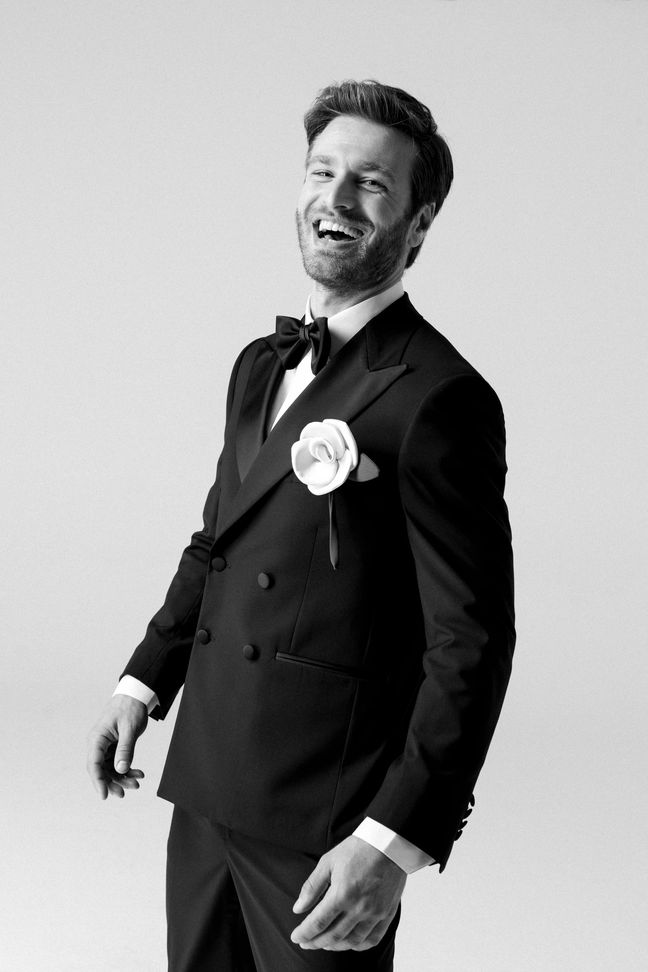 Uomo sorridente in abito da sera con fiore sulla giacca, in bianco e nero.