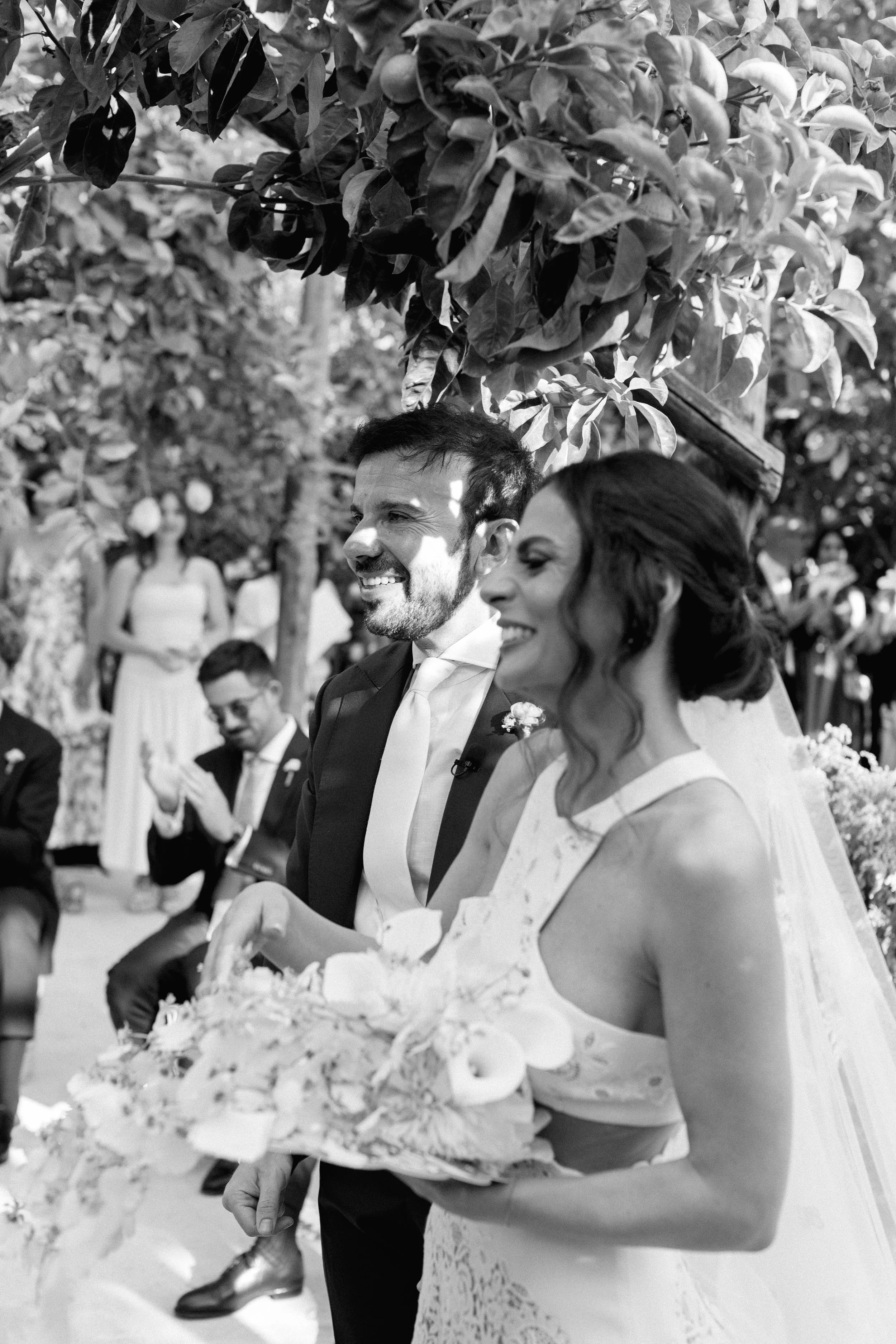 Una coppia di sposi durante il matrimonio, sorridenti, circondati da altri invitati seduti, sotto un albero con foglie.