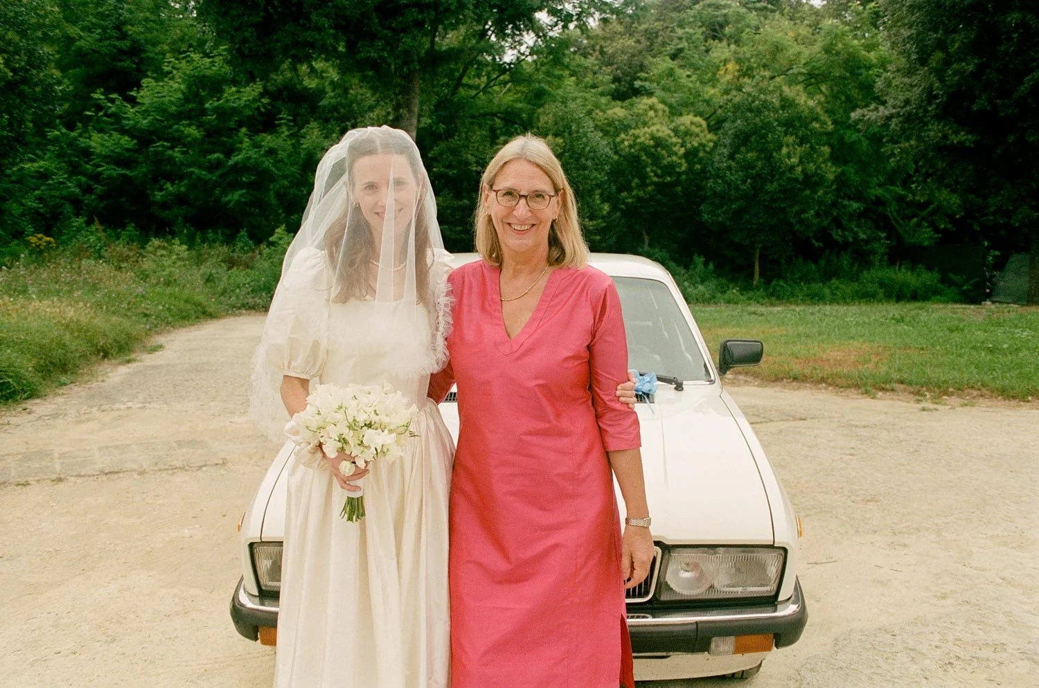 Donna in abito da sposa con velo e bouquet vicino a una donna con abito rosa, davanti a un'auto bianca, in un'area con alberi e natura.