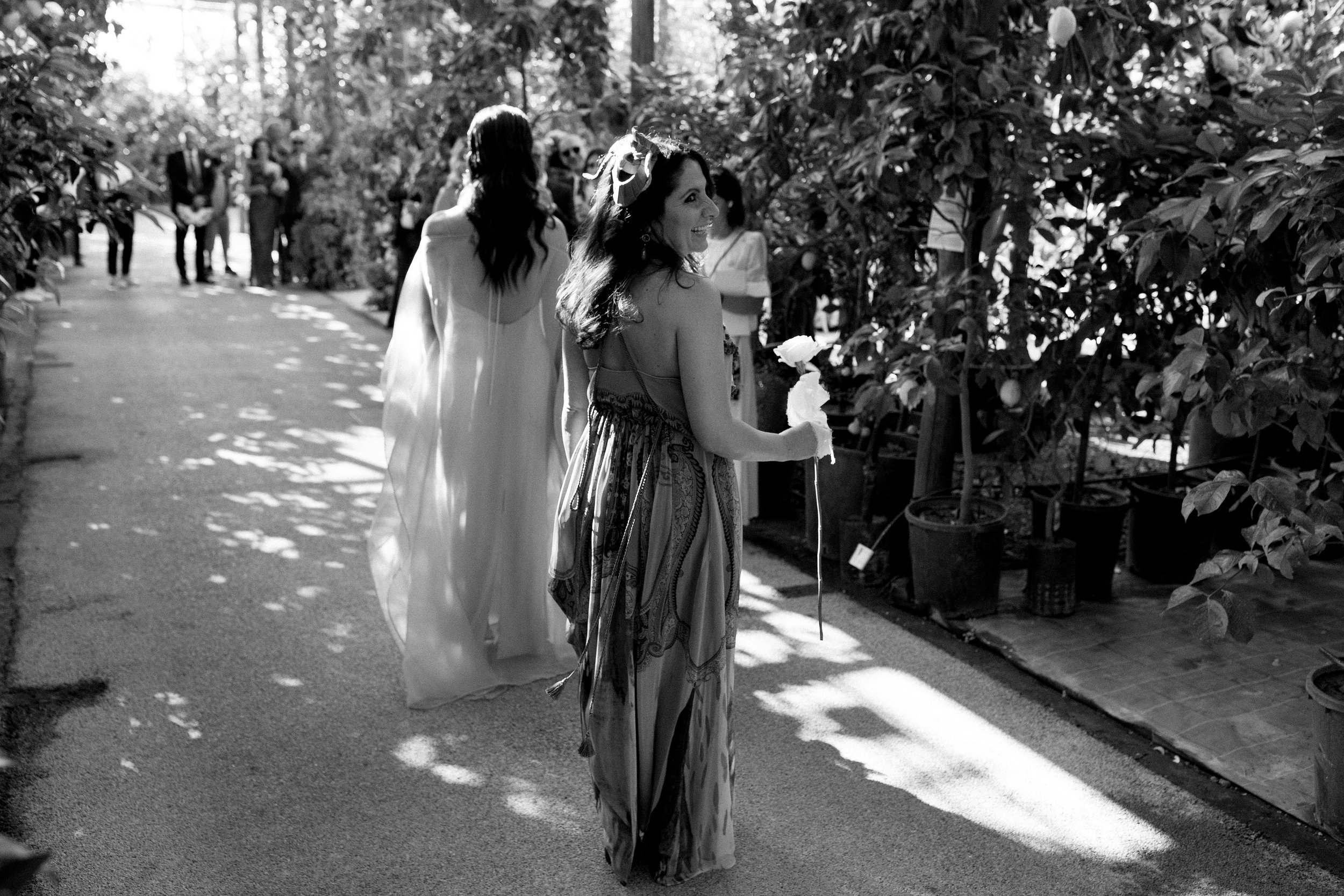 Donne che partecipano a un matrimonio o a un evento in un ambiente all'aperto, alcune con abiti lunghi, in penombra con alcune aree illuminate dal sole, piante e vasi sullo sfondo.