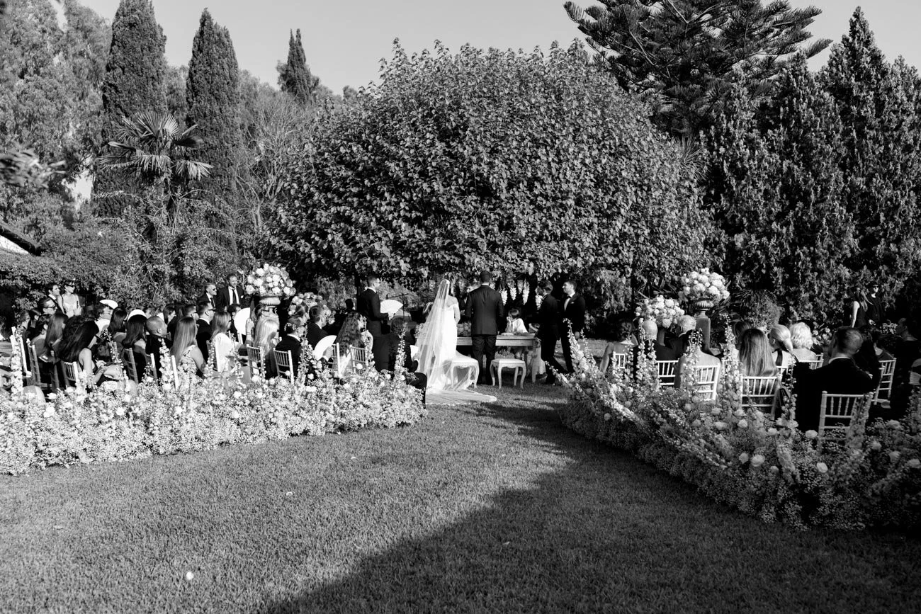 Un matrimonio si svolge in un giardino durante il giorno, con invitati seduti su sedie intorno a un tavolo, decorato con fiori. Gli sposi sono in piedi davanti a un cerimoniere, con molti alberi e foliage sullo sfondo.