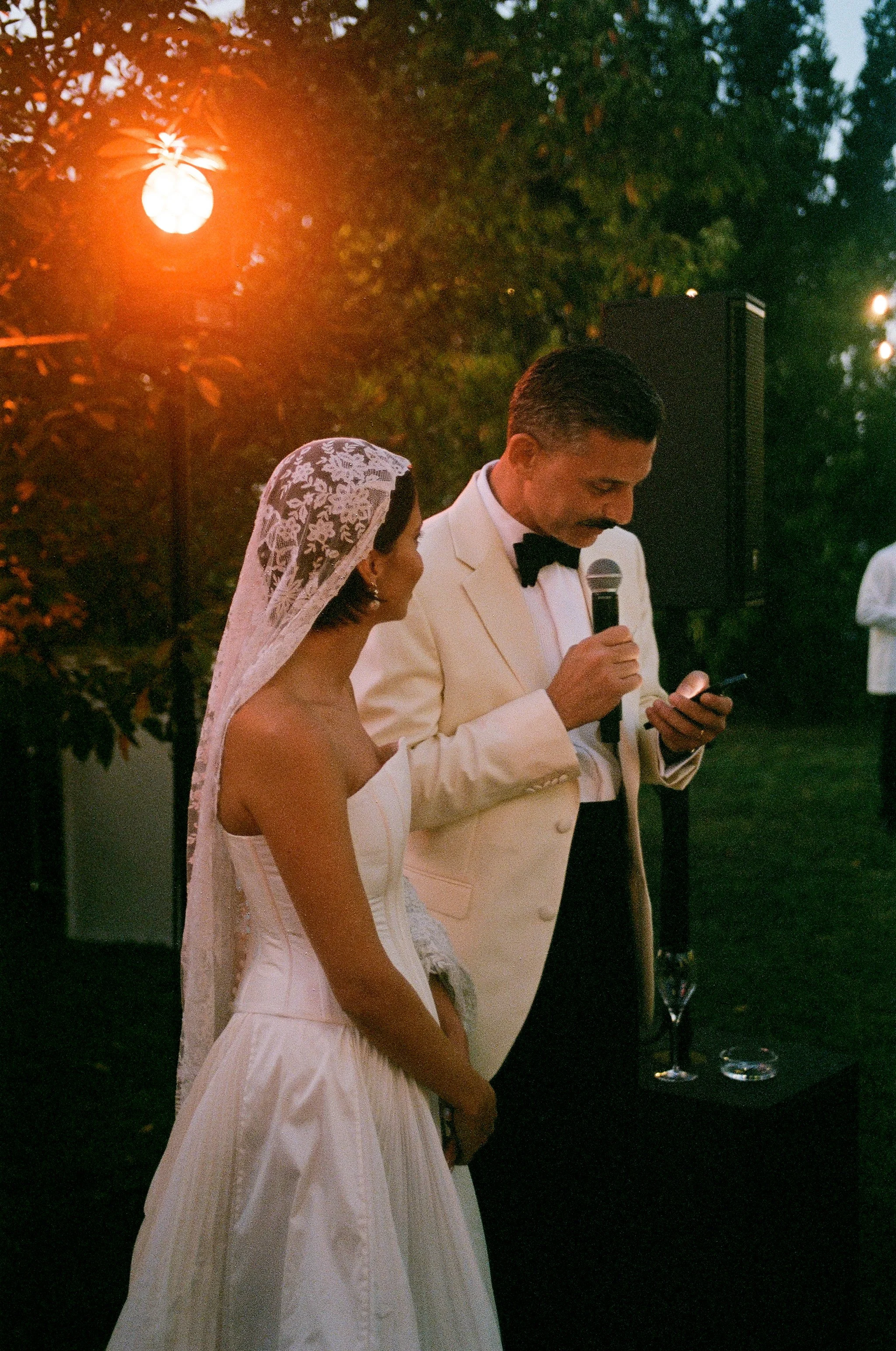 Due persone vestite elegantemente durante un matrimonio all'aperto, con alberi e luci calde sullo sfondo.