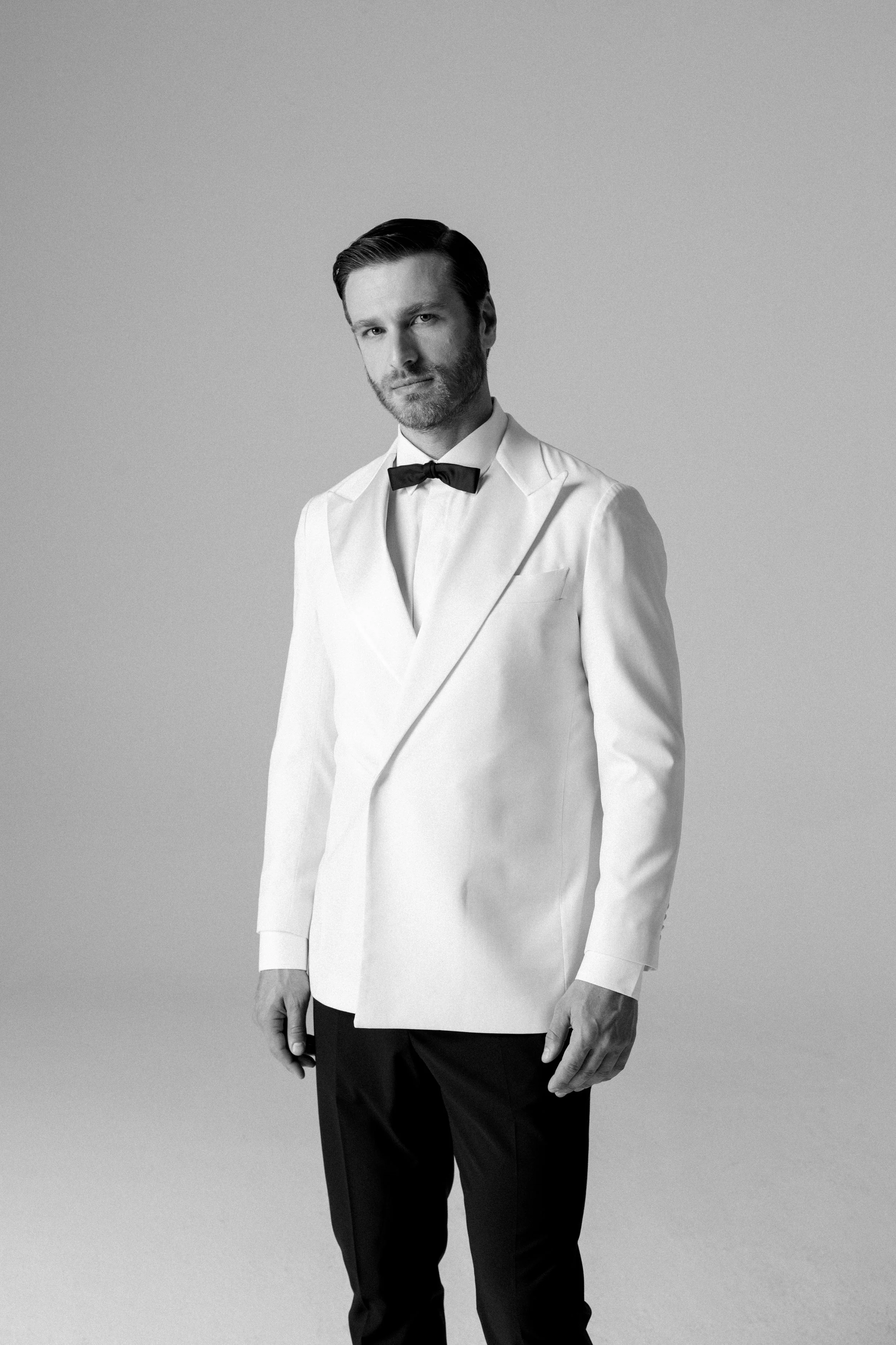 Uomo elegante con abito bianco e papillon nero davanti a sfondo neutro.