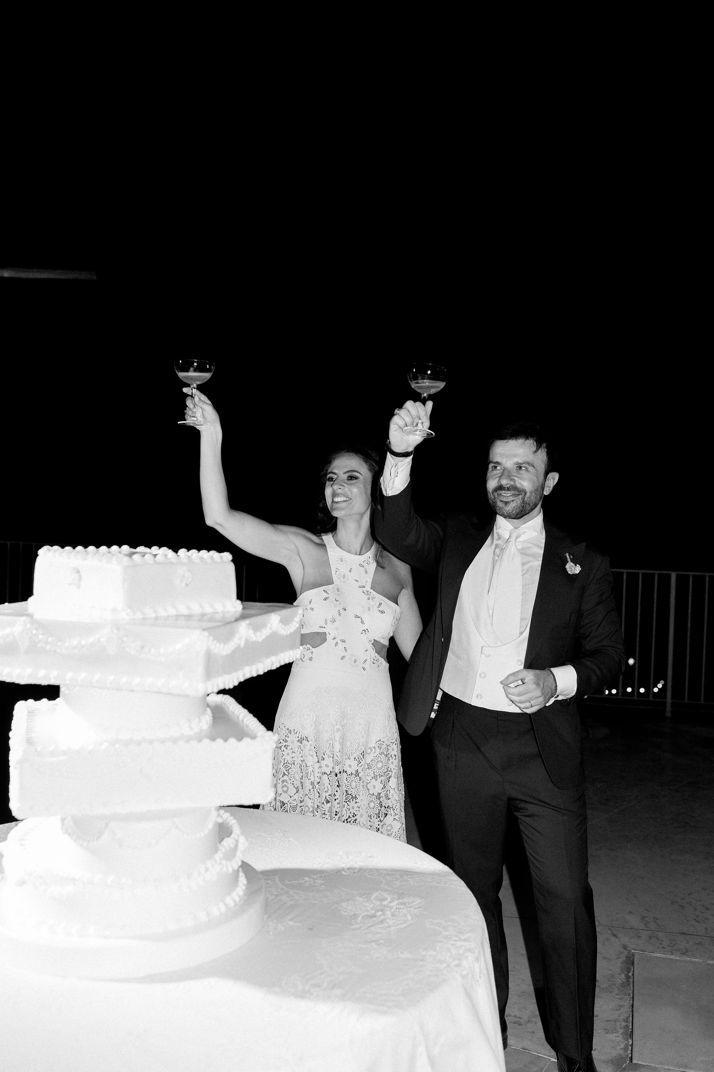 Una coppia di sposi brinda con bicchieri di vino durante il loro matrimonio, con una torta nuziale in primo piano.