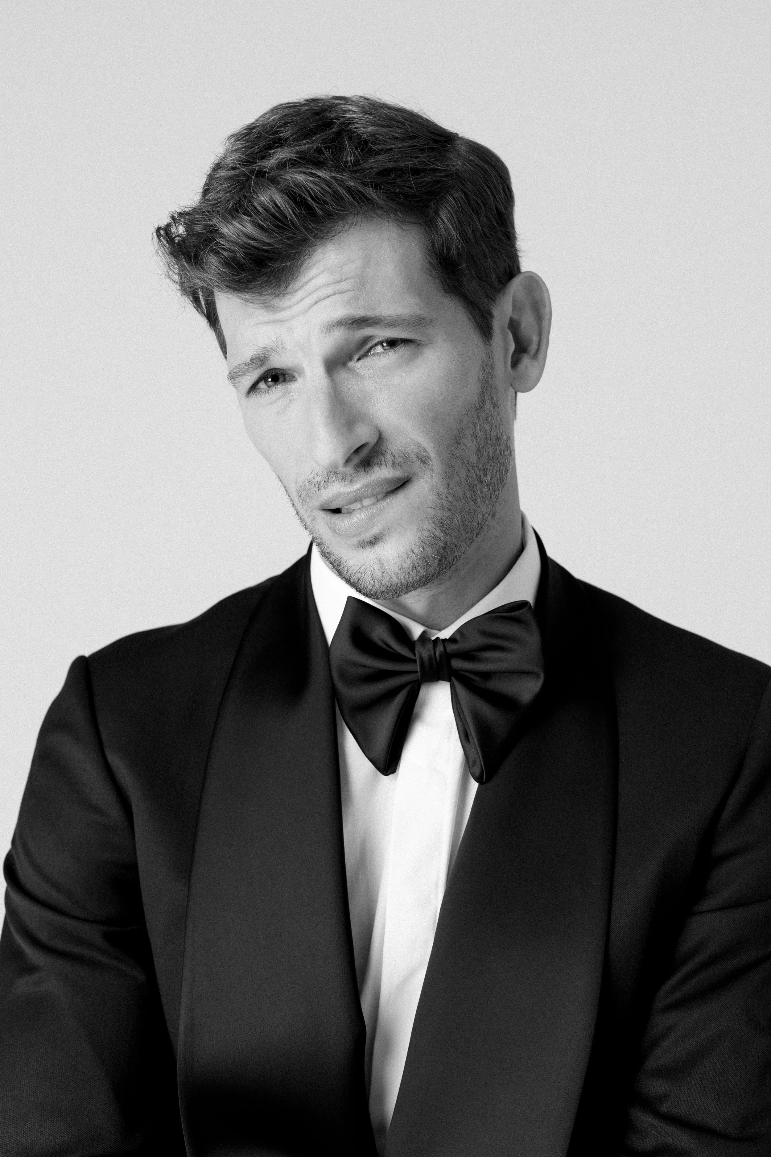 Ritratto di un uomo giovane con capelli castani, con un'espressione confusa, indossa un abito elegante nero e una cravatta a farfalla, sfondo chiaro.