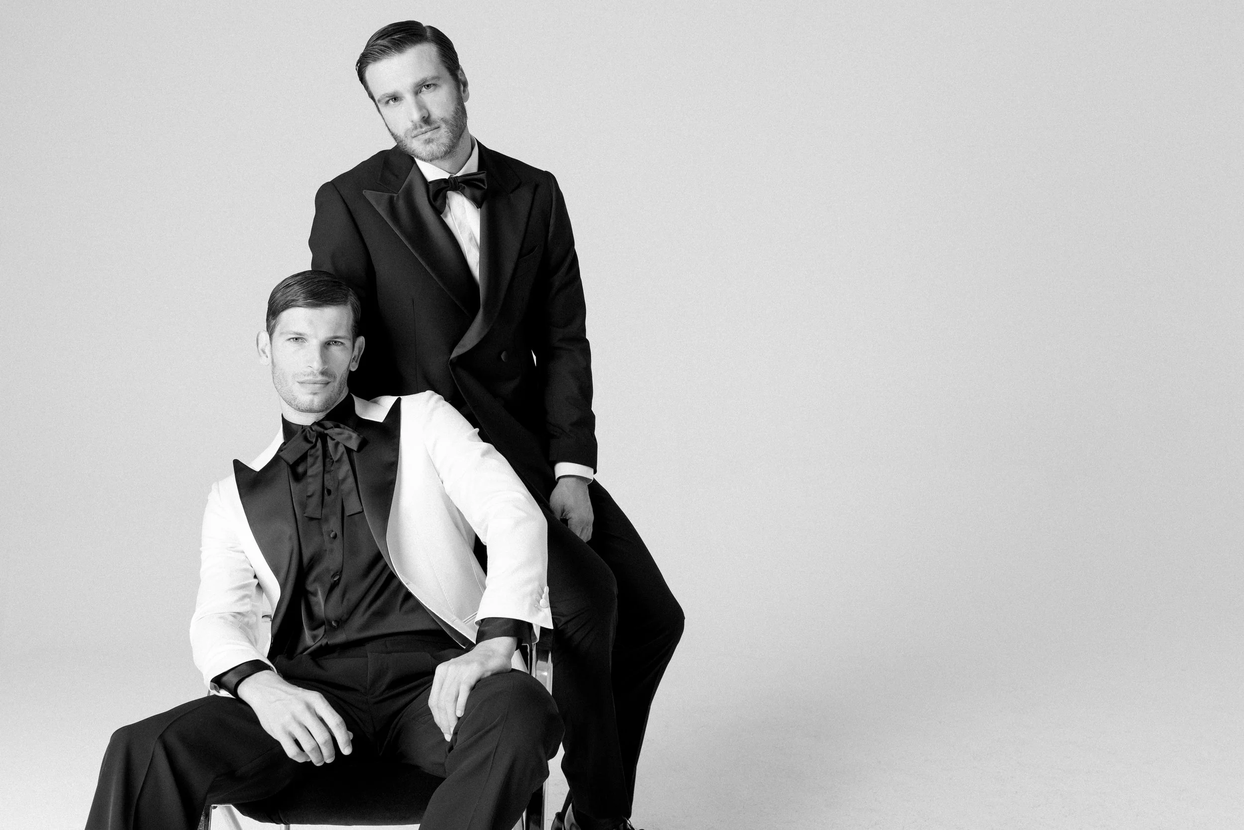 Due uomini eleganti in abiti da sera neri e bianchi con papillon, uno seduto e uno in piedi, in uno studio fotografico a colori o in bianco e nero.