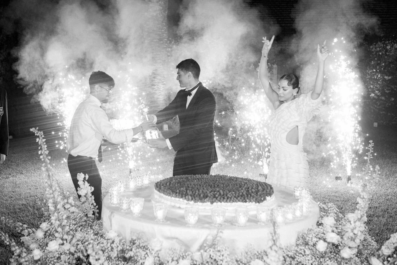 Una scena di celebrazione di nozze con due persone, una in abito da sposo e l'altra in abito scuro, che si scambiano un regalo davanti a una torta decorata, circondati da fuochi d'artificio e fumo, con una donna in abito elegante che ringrazia o fest