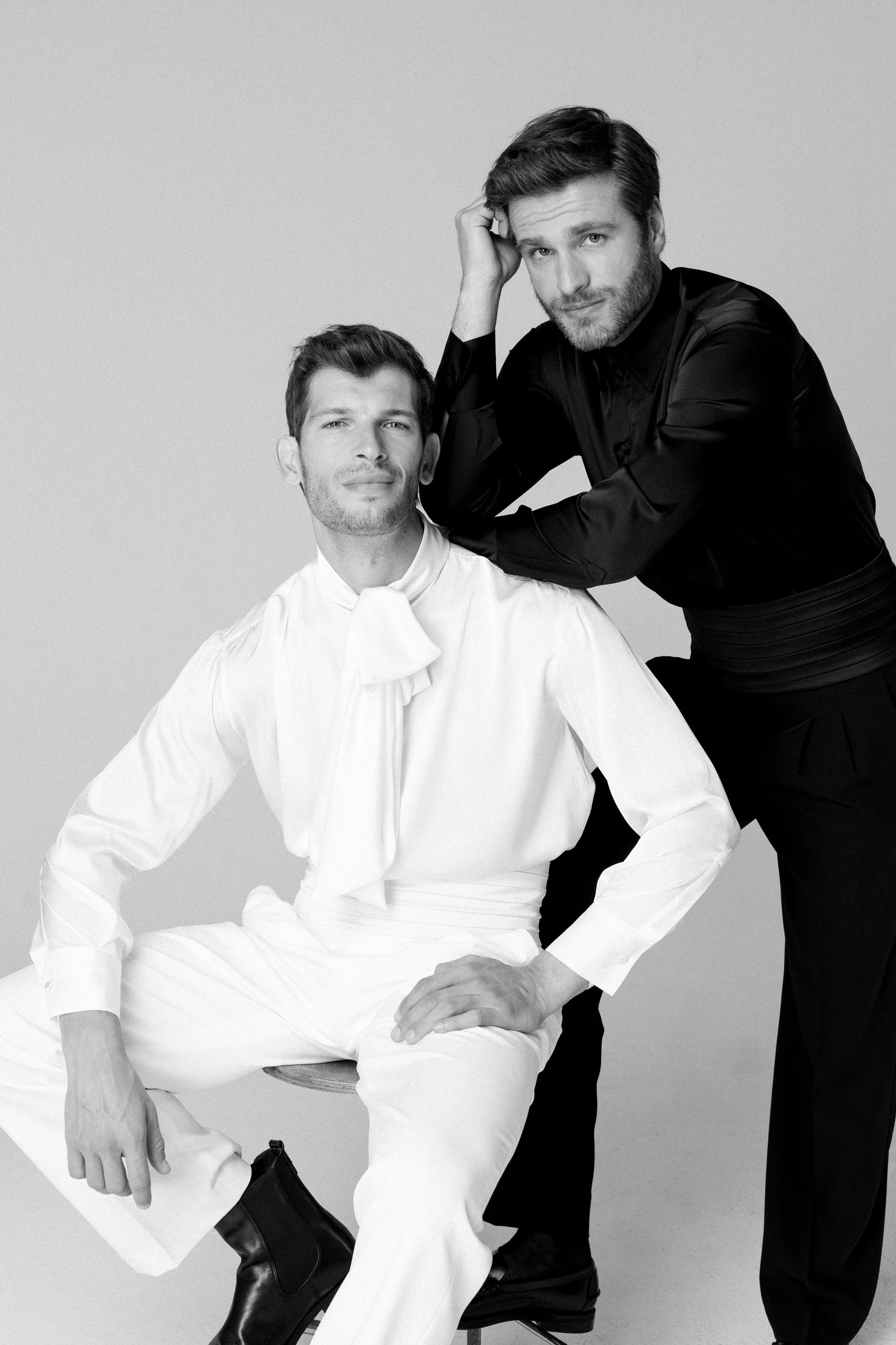 Due uomini in bianco e nero; uno seduto e uno in piedi, entrambi elegantemente vestiti.