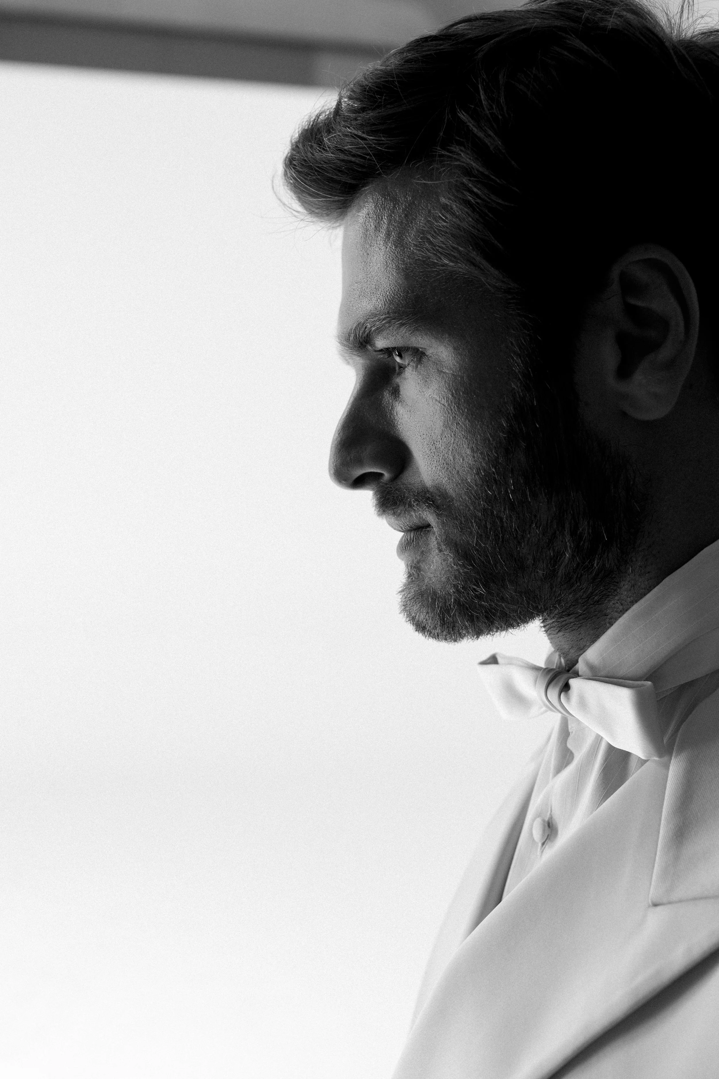 Ritratto in bianco e nero di un uomo sofisticato con barba, capelli castani e abiti eleganti, incluse giacca e papillon, osservando lateralmente.