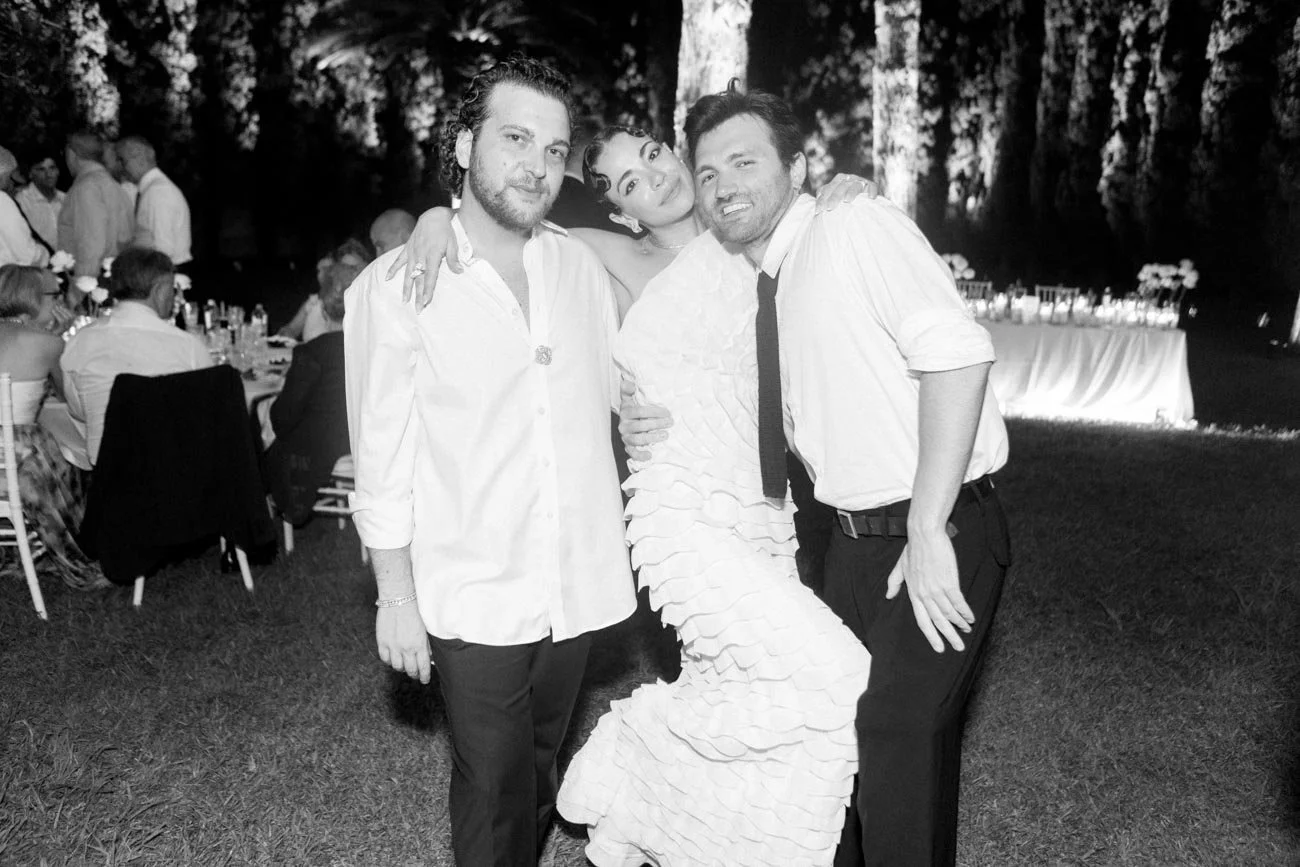 Tre persone sorridenti durante un evento, con due uomini e una donna in abiti eleganti, posano per la foto su un prato con tavoli e persone sedute sullo sfondo.