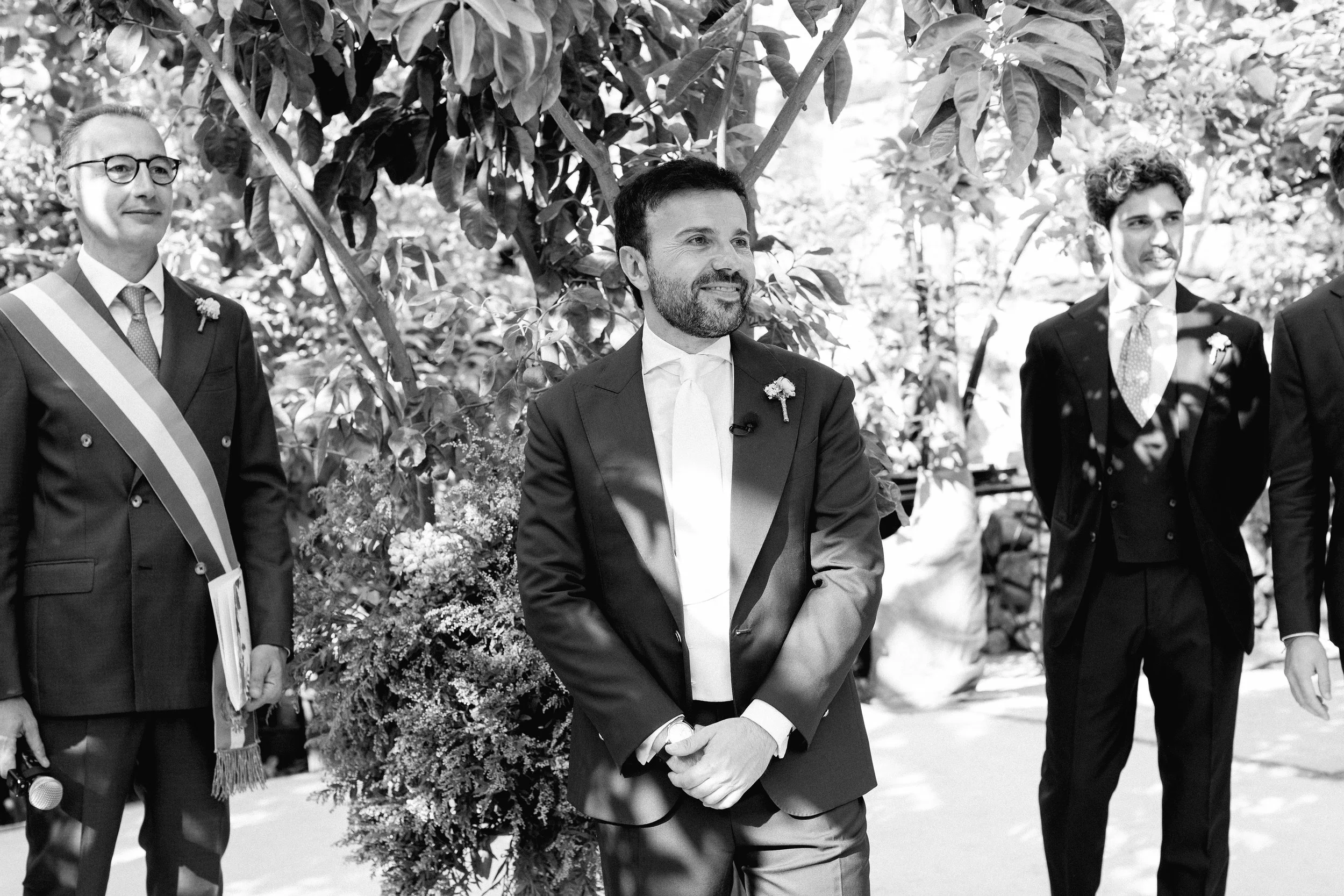 Un gruppo di uomini in abiti formali durante una cerimonia all'aperto, con uno sfondo di piante e fiori.