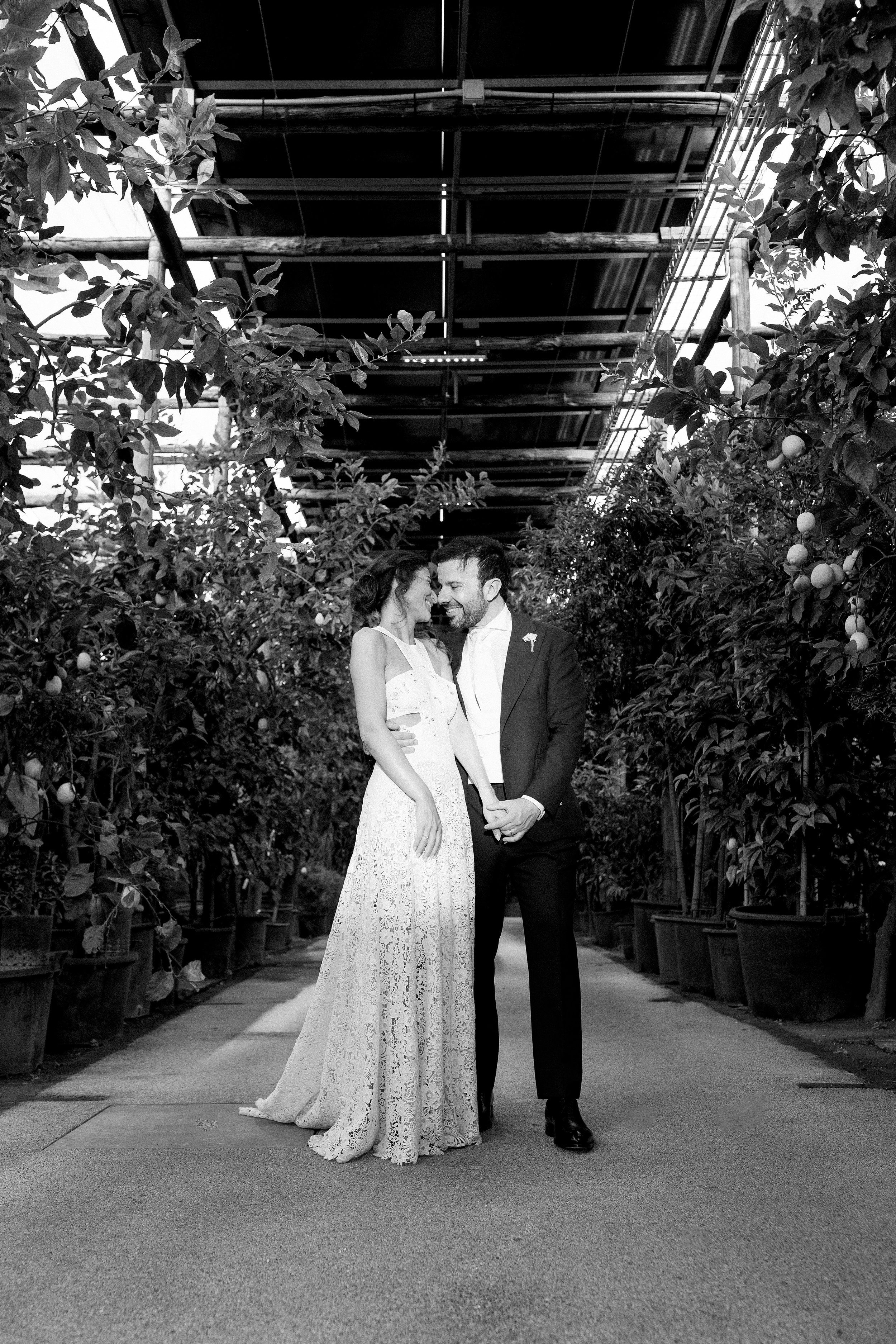 Una coppia di sposi si abbraccia e sorride in un ambiente con piante di arance e un soffitto in metallo, in bianco e nero.