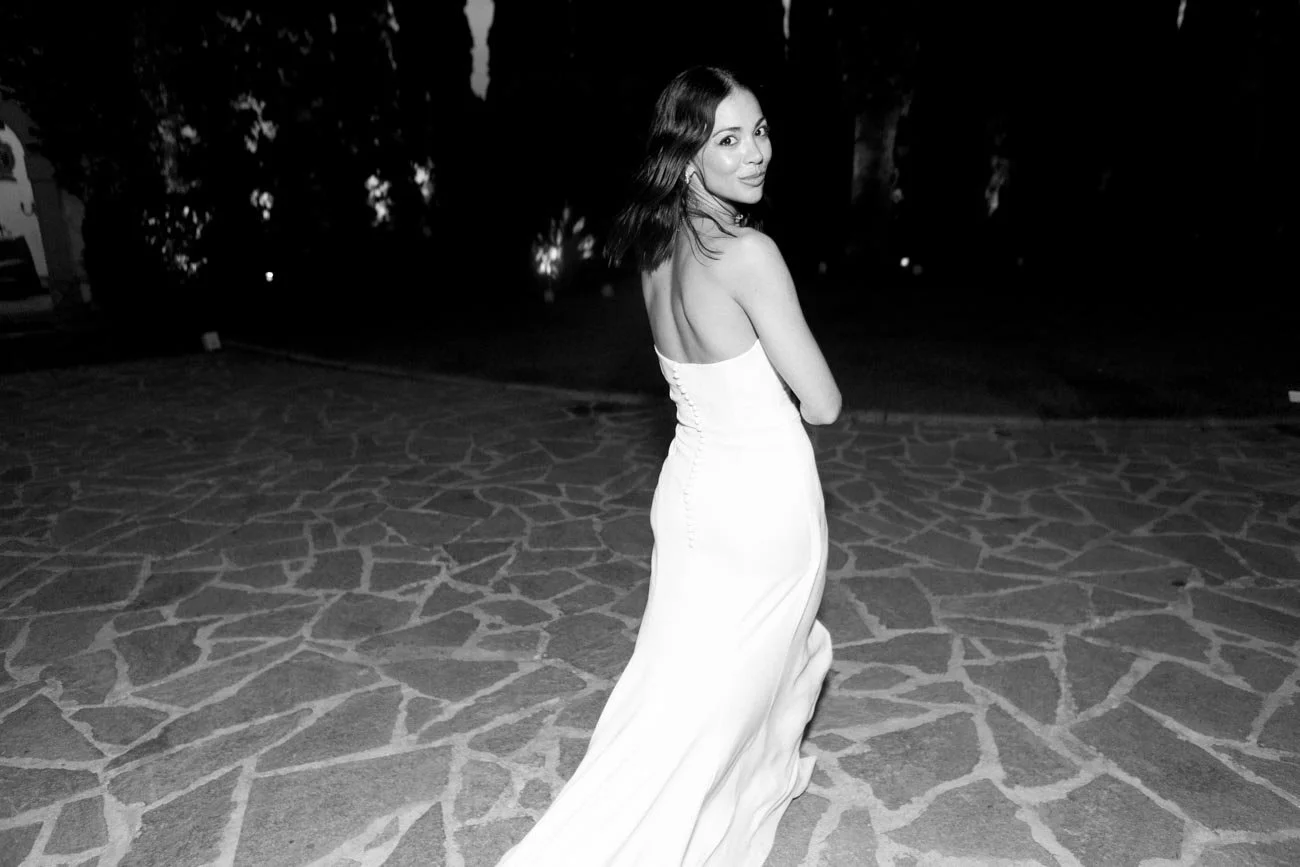 Donna in abito lungo bianco, con capelli castani, che si gira e sorride, in un ambiente all'aperto con pavimento in pietra e alberi sullo sfondo, in bianco e nero.
