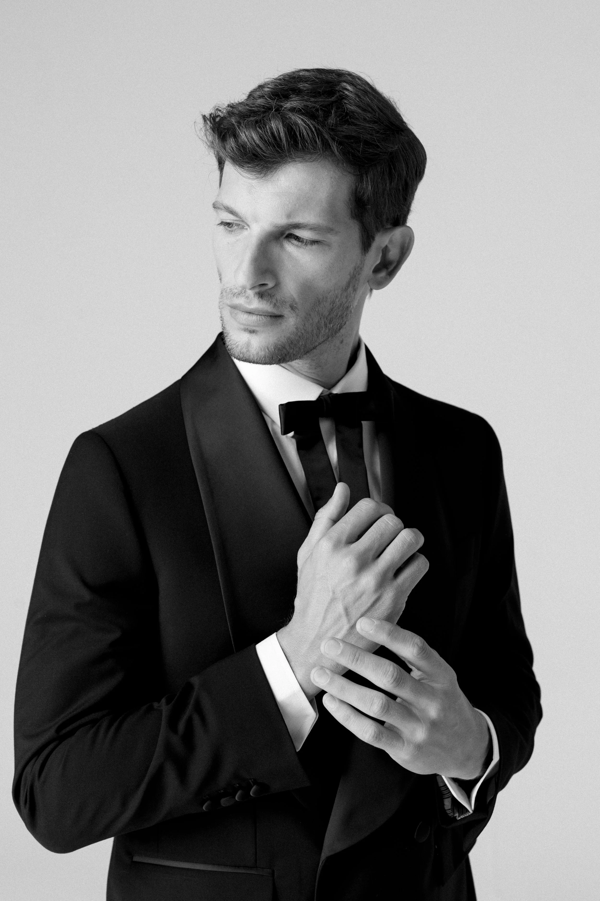 Un uomo in abito elegante con fermaglio a farfalla, posa con le mani avvolte e si guarda di lato contro uno sfondo chiaro.