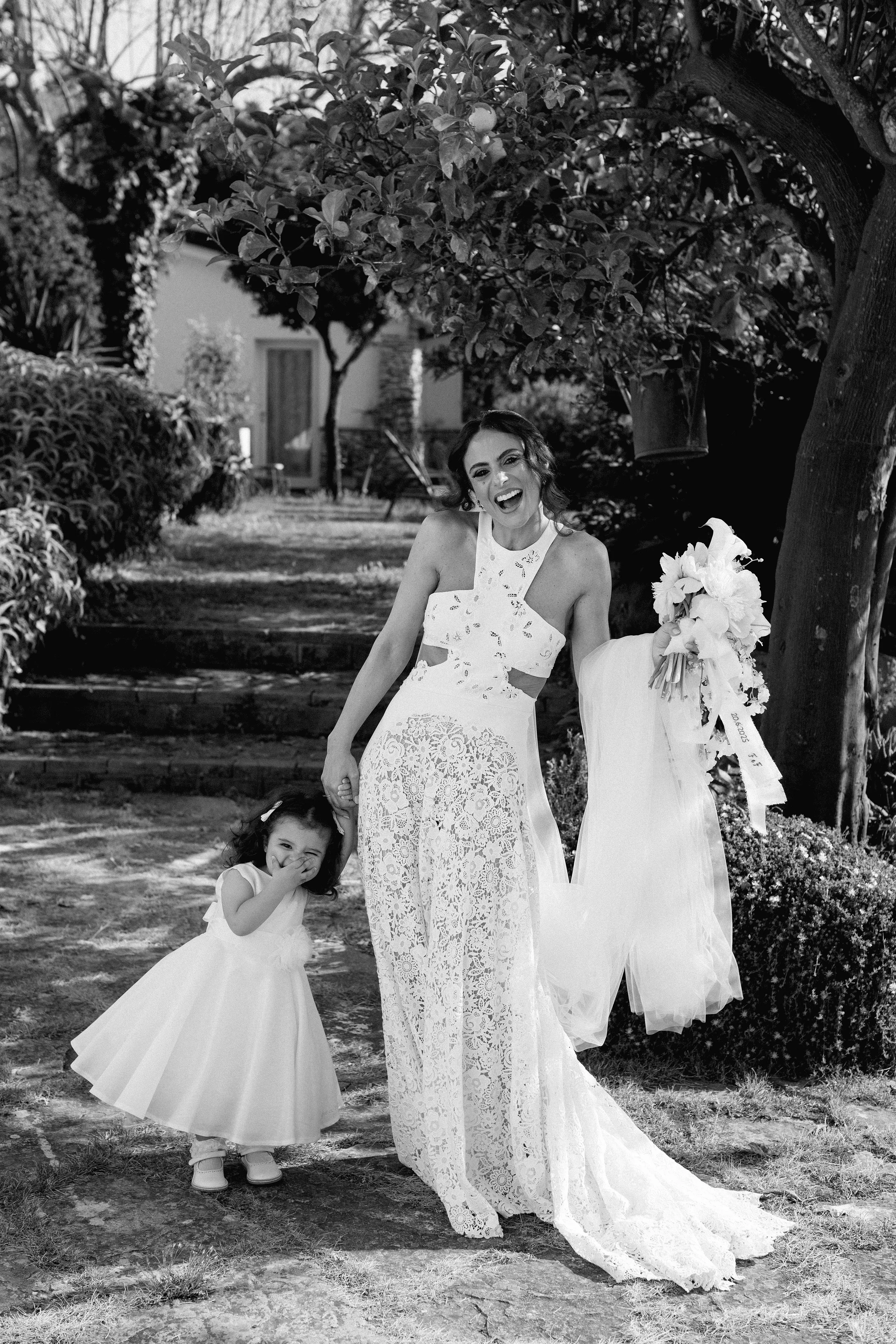 Una donna sorridente con un vestito da matrimonio accanto a una bambina in un abito bianco in un giardino