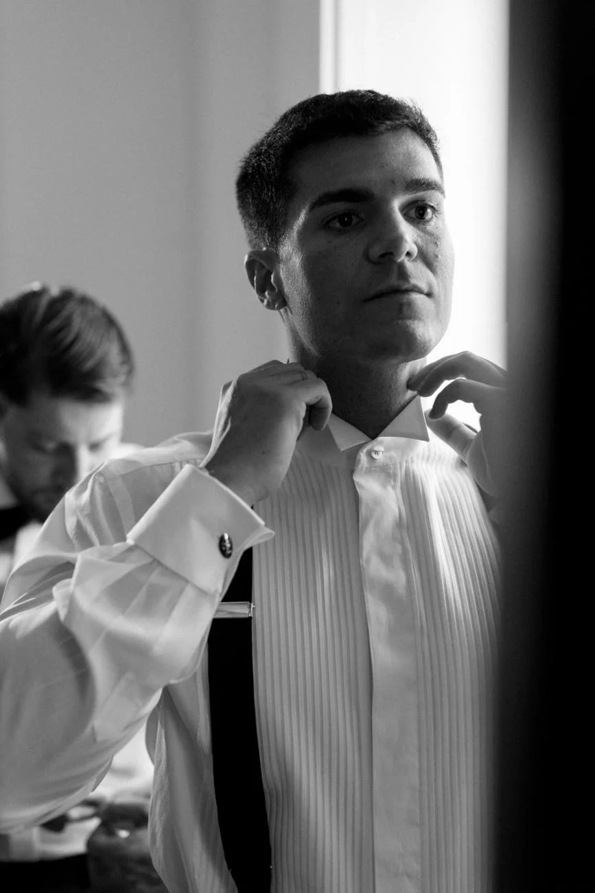 Un uomo mentre si sistema il colletto della camicia, un uomo e una donna al bakcground, sono in un ambiente intimo e silenzioso.