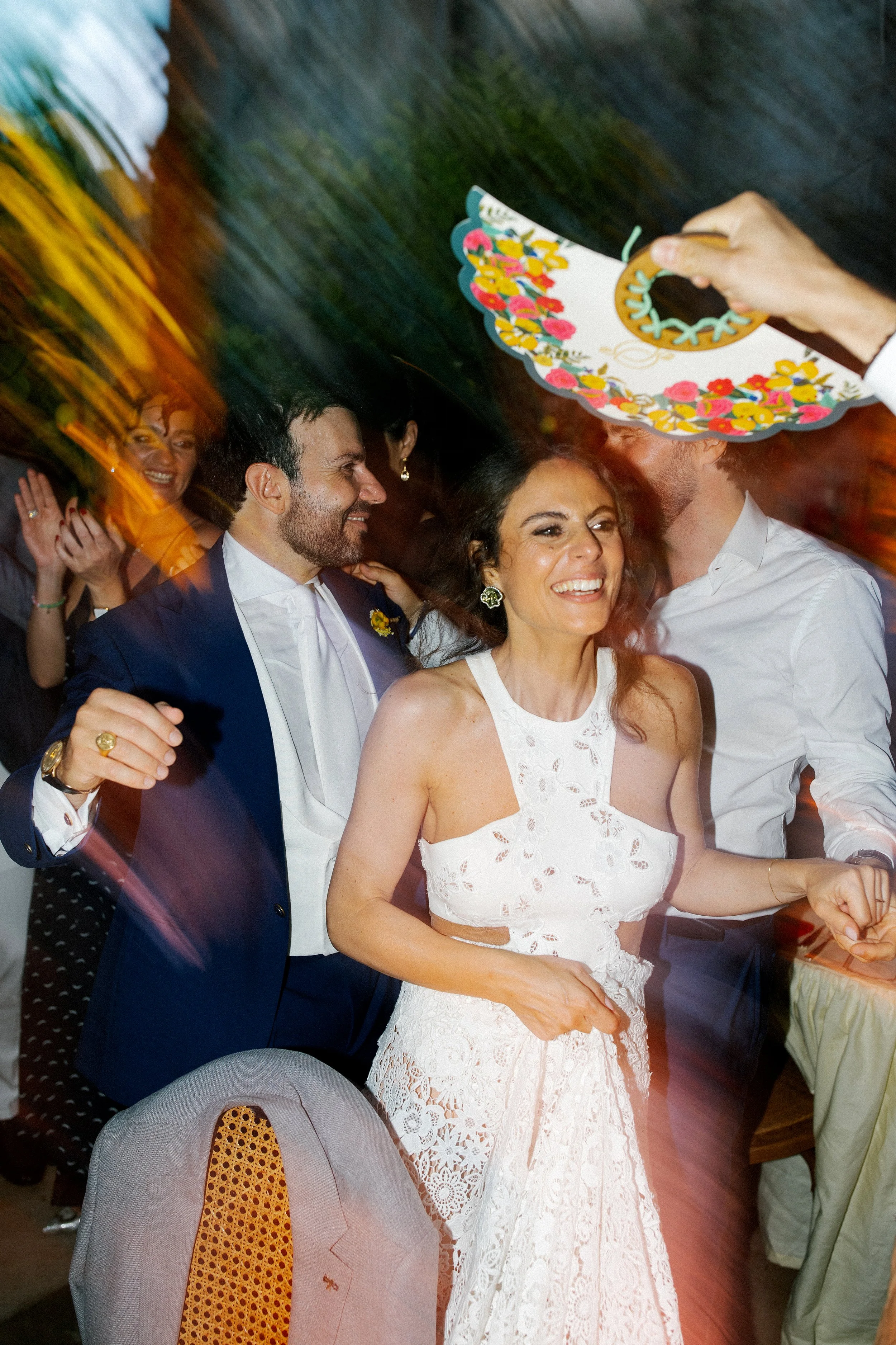 Coppia felice durante un matrimonio. La donna sorride e indossa un vestito bianco in pizzo, mentre l'uomo la guarda con affetto. Qualcuno le sta consegnando un simbolico anello decorato con fiori.