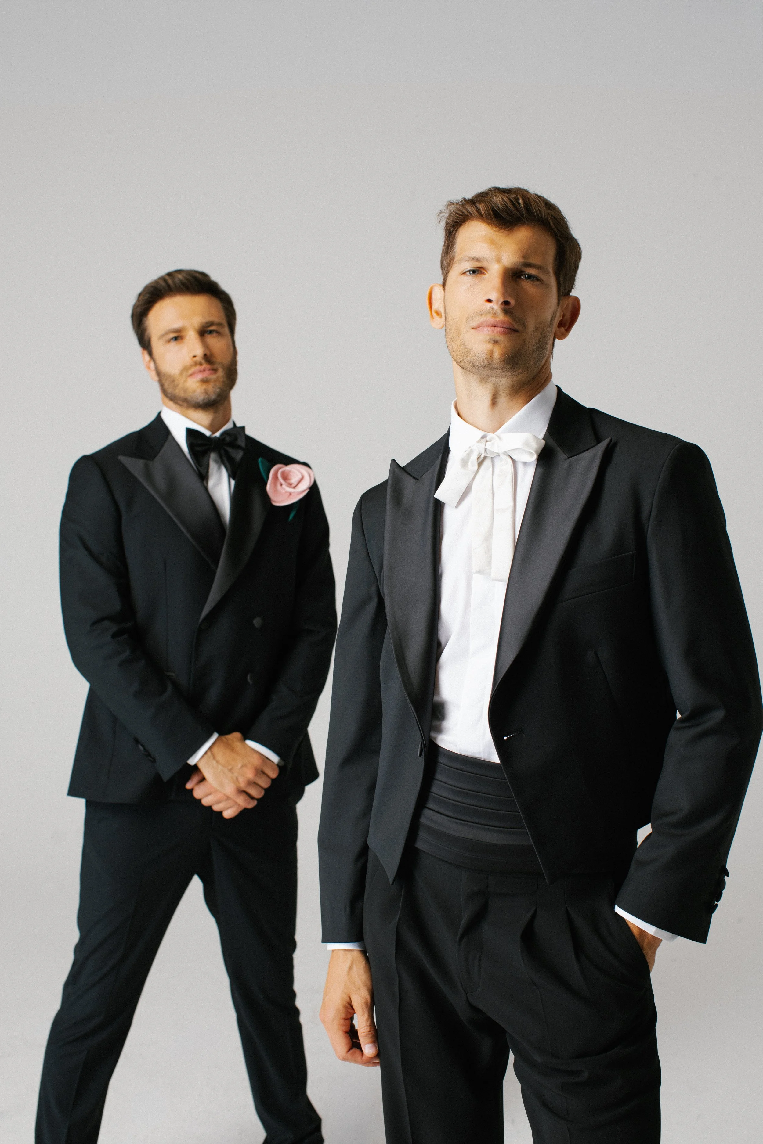 Due uomini in abiti formali neri con camicie bianche e cravatte, uno indossa un papillon, si trovano su uno sfondo neutro grigio, uno davanti e uno dietro.