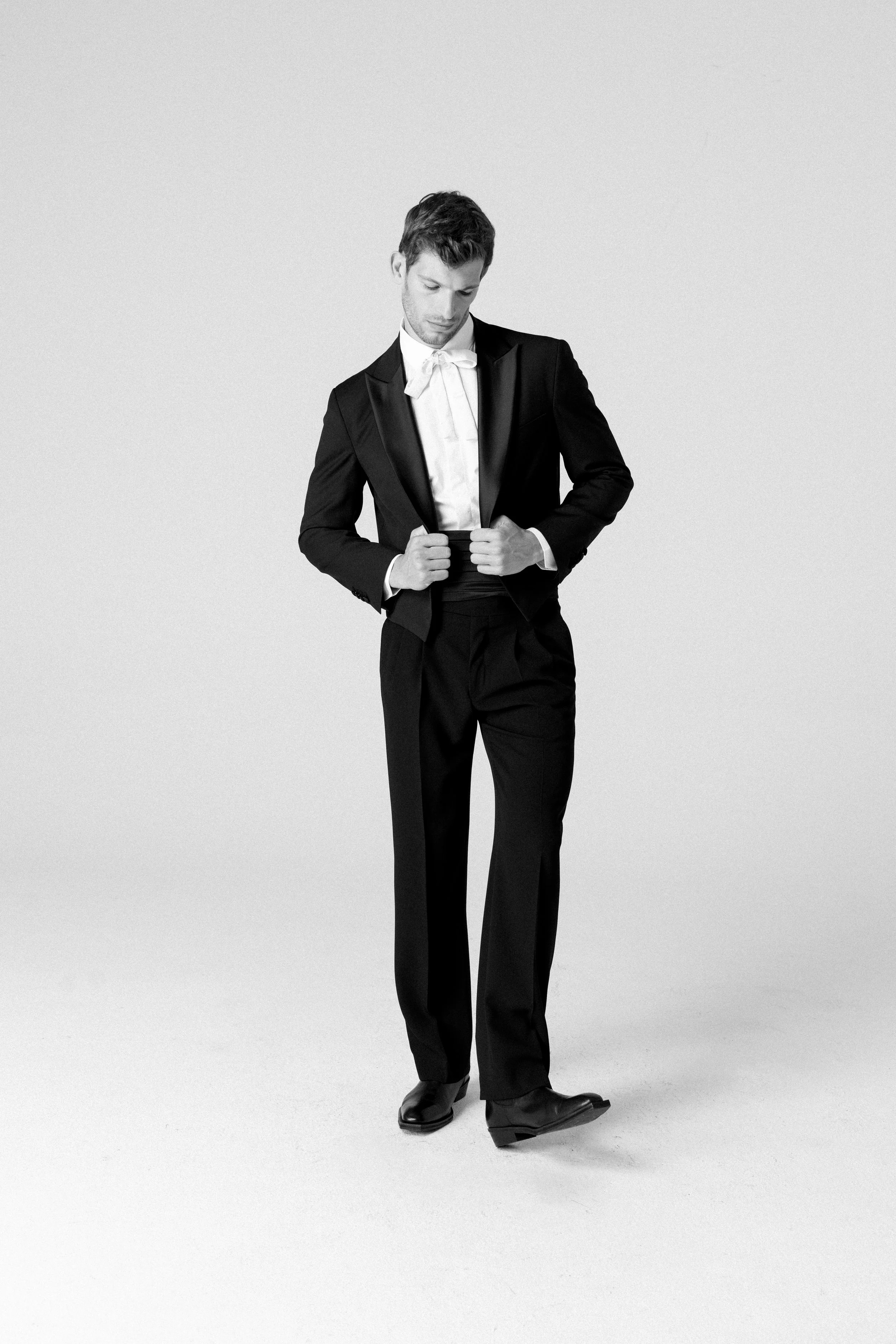 Uomo con abbigliamento formale nero, giacca e pantaloni, posa in piedi con le mani sui bottoni della giacca, sfondo bianco.