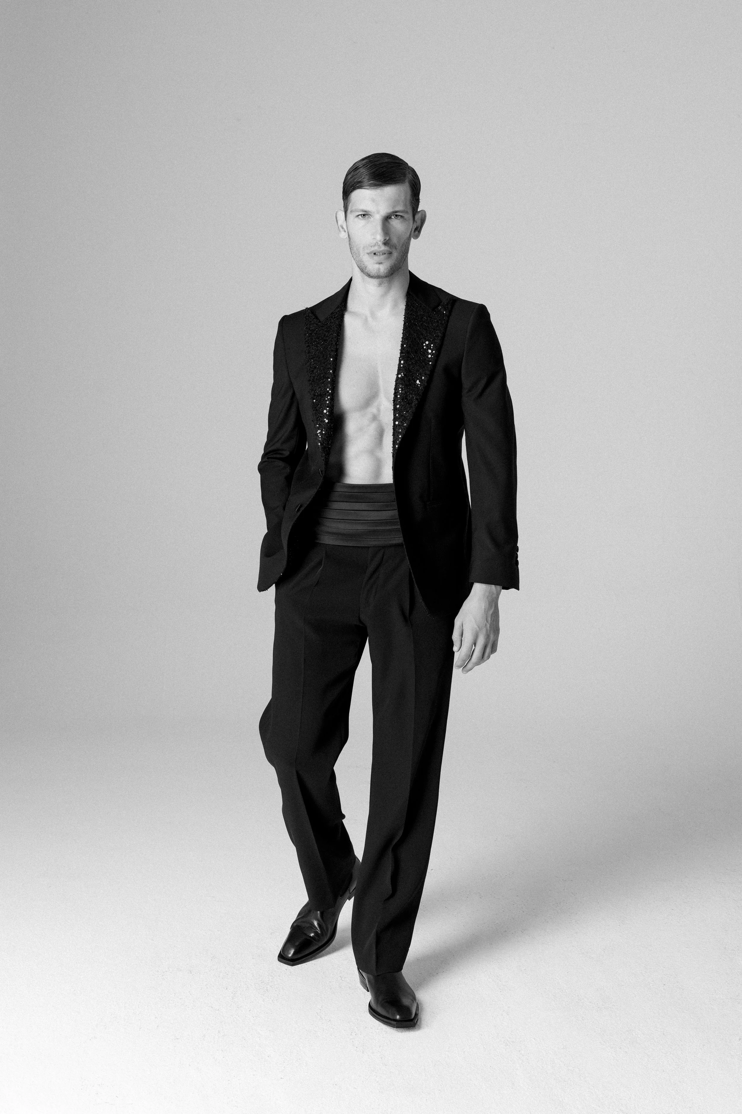 Uomo in abito nero con giacca aperta e senza camicia, in posa in un ambiente neutro.