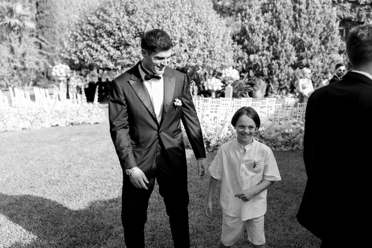 Un ragazzo vestito elegantemente con un abito nero e una cravatta a farfalla sorride mentre cammina all'aperto con un bambino che indossa una camicia bianca e pantaloncini, in un giardino durante un evento formale.