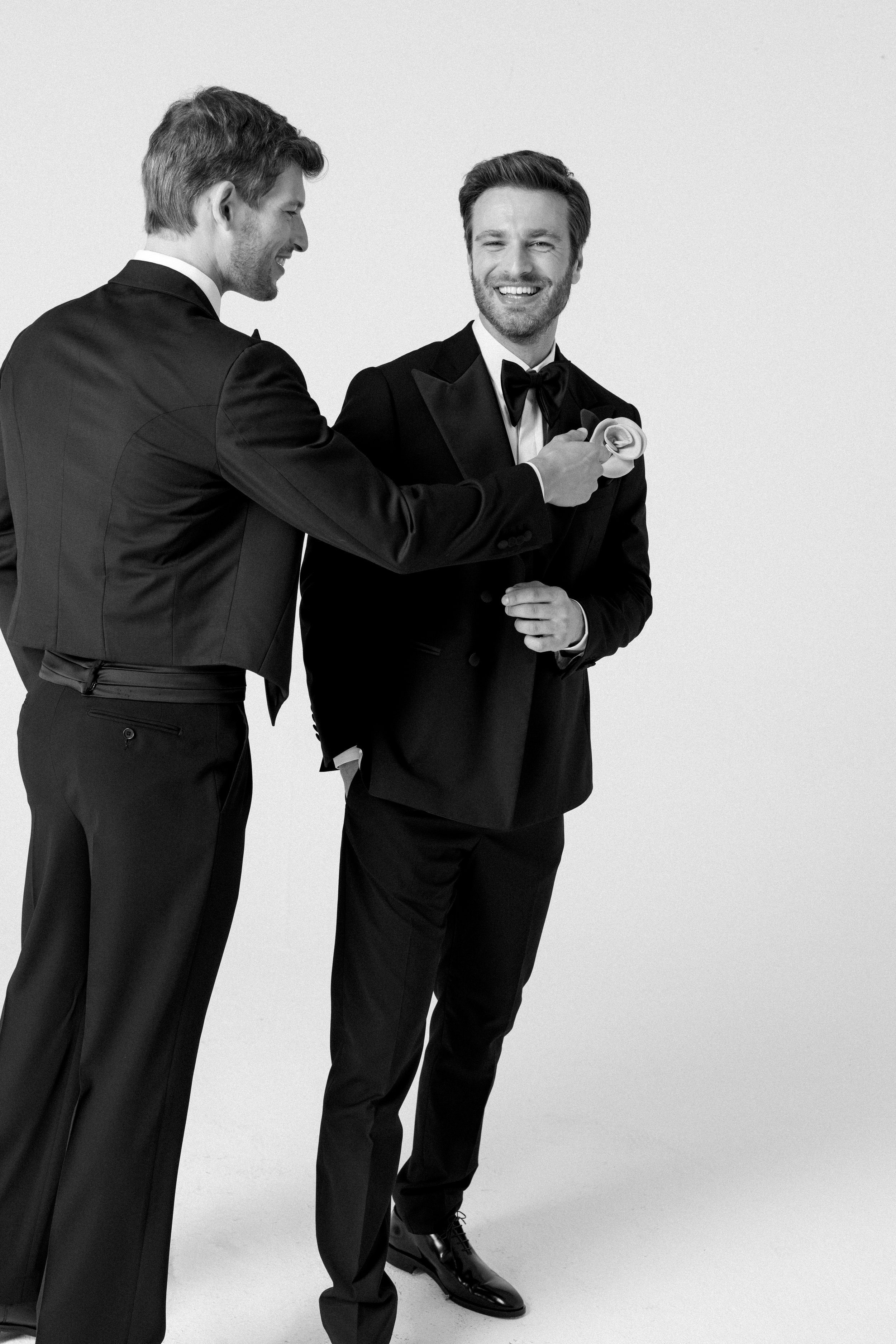 Due uomini vestiti con smoking eleganti sorridono e uno dei due tiene un fiore in mano