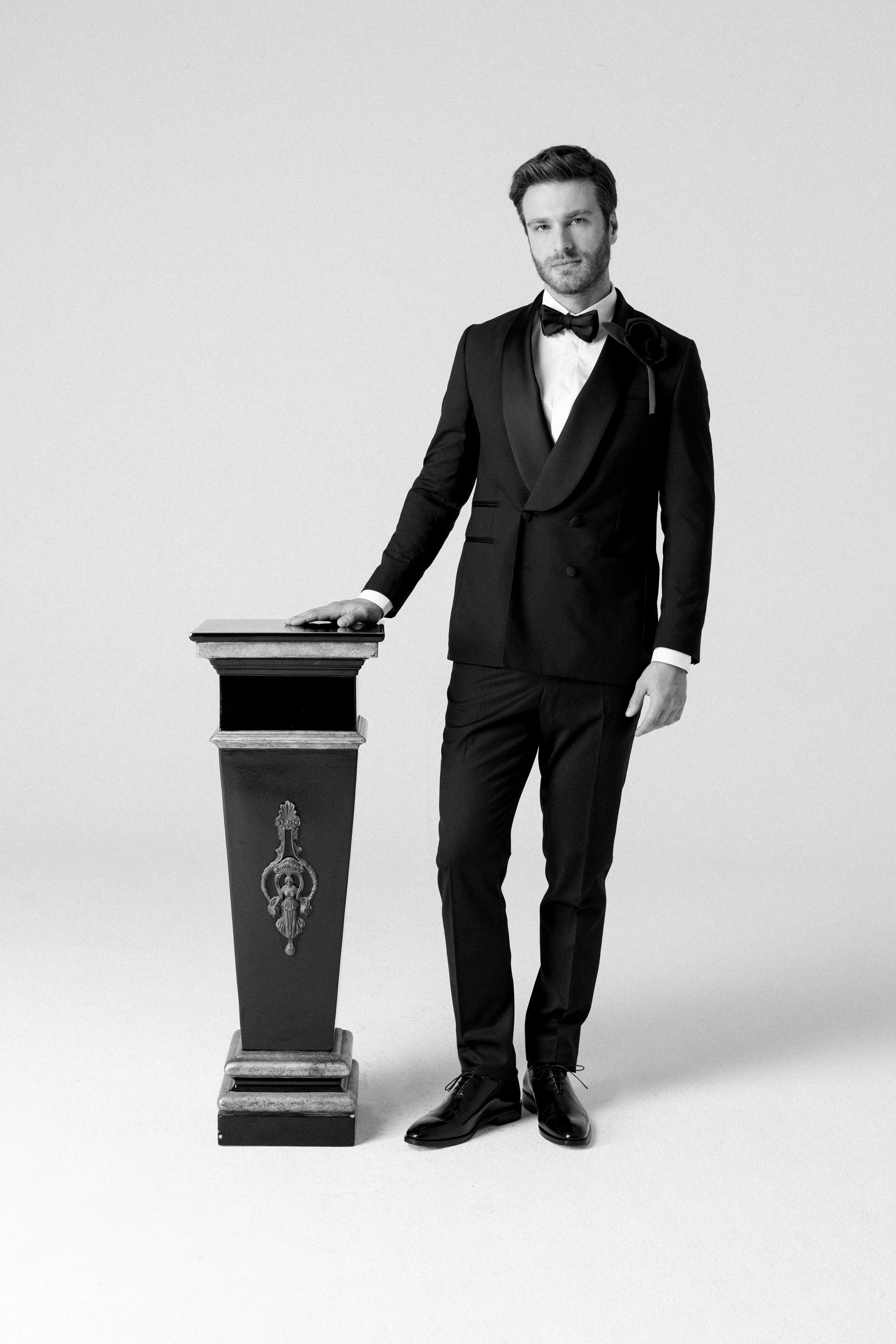 Uomo in completo da cerimonia nero con papillon, che posa con una mano su un podio decorato, contro uno sfondo bianco.