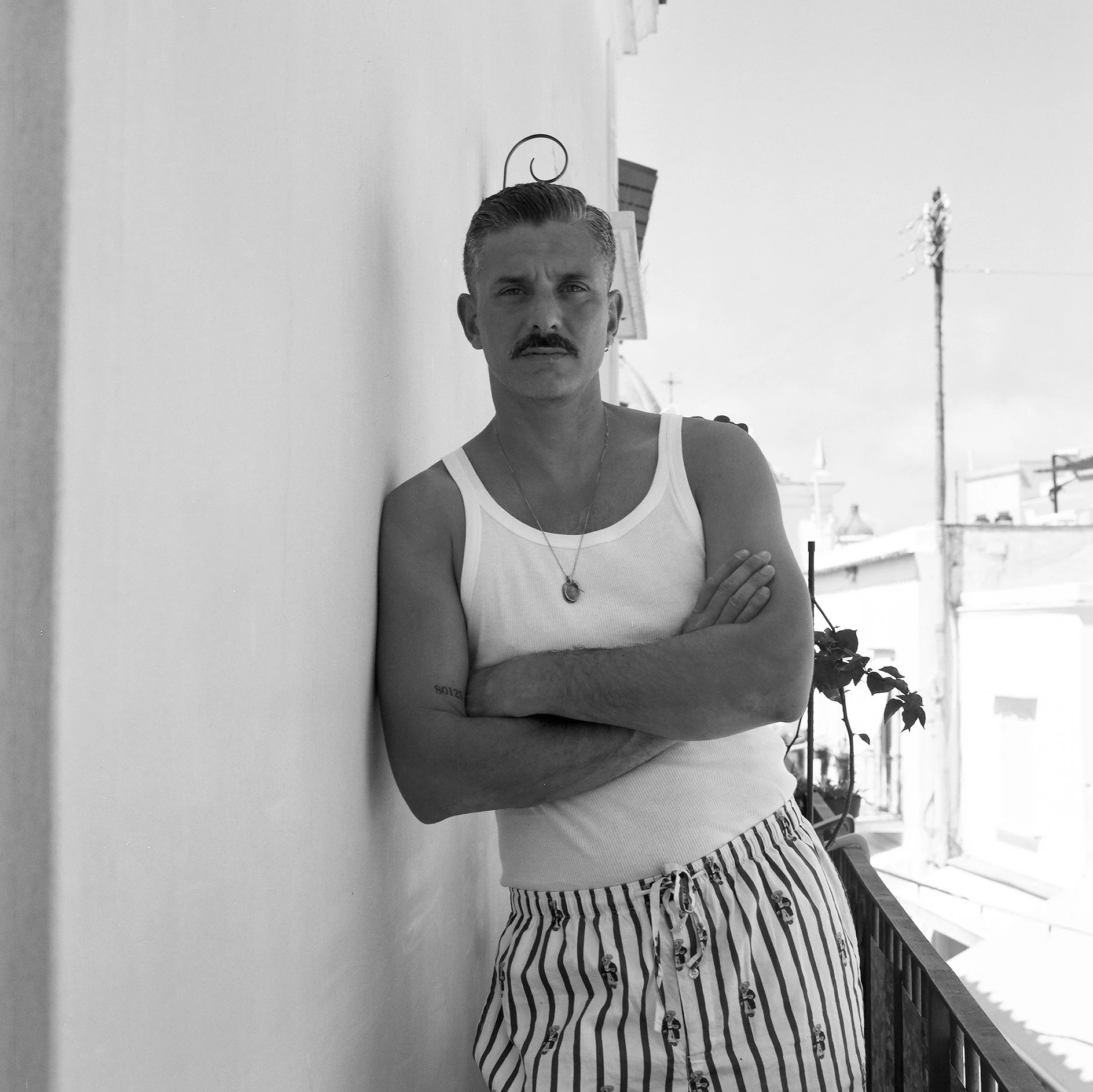 Uomo con baffi e cappello, vestito con canottiera bianca e pantaloni a righe, si appoggia a un muro di fronte a un balcone, con le braccia incrociate.