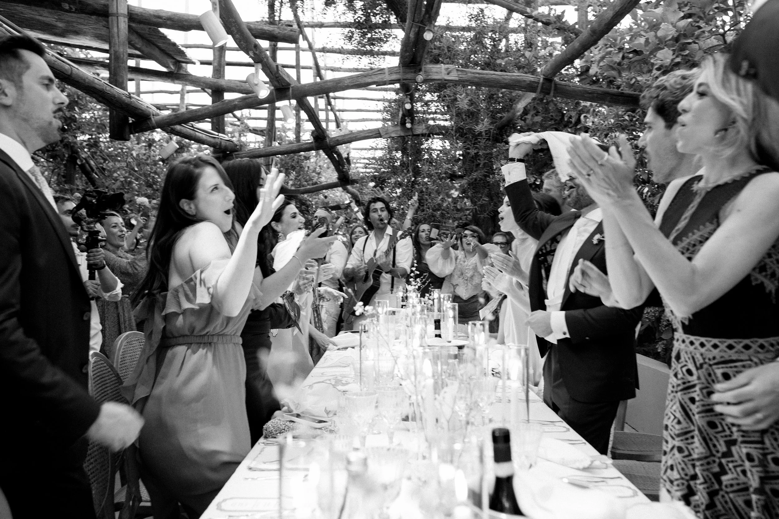 Un gruppo di persone a un matrimonio si sta divertendo, alcune cantano e altre ballano attorno a un tavolo con candele e bottiglie, in un ambiente con un soffitto di legno e luci