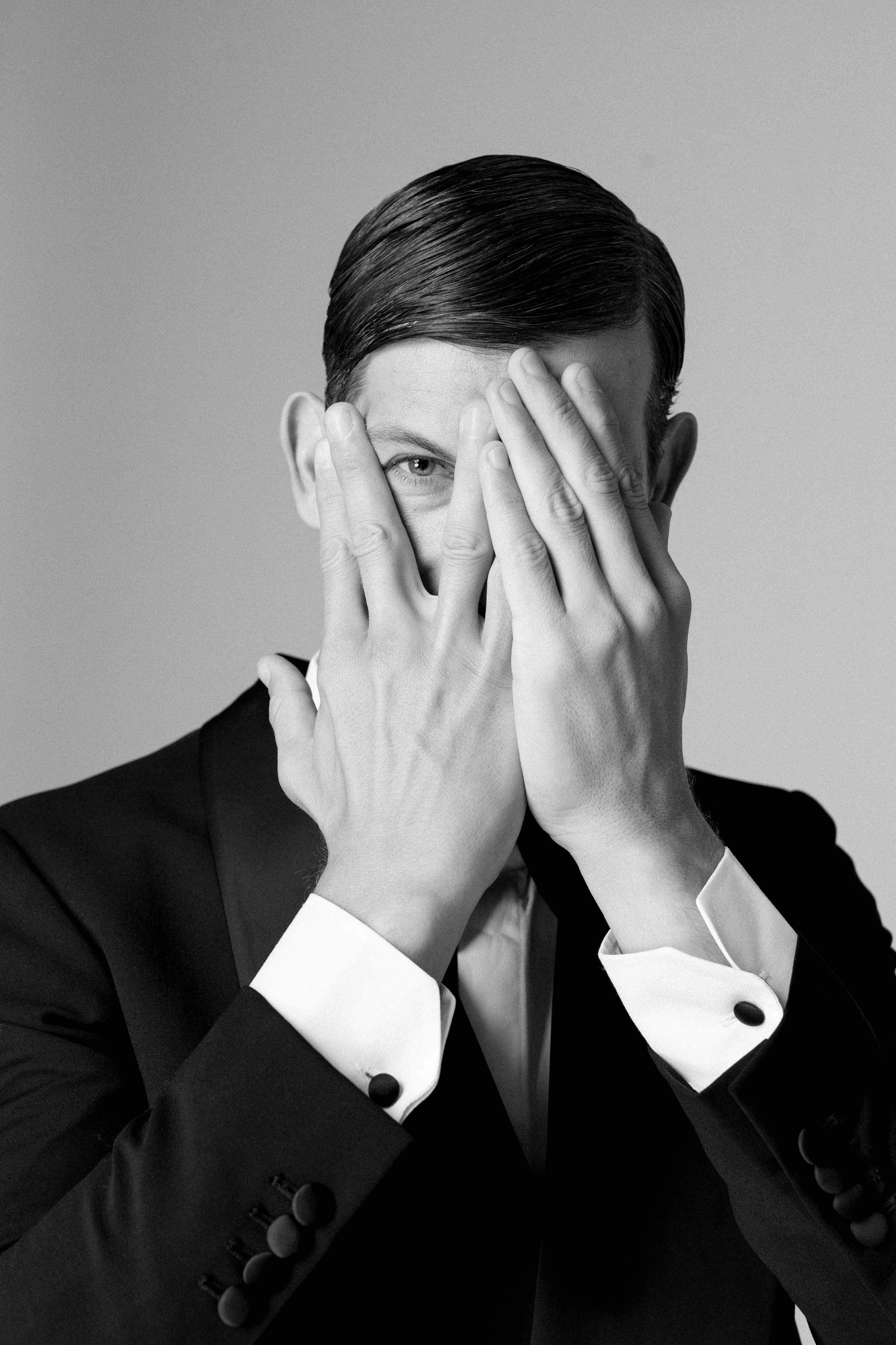 Ragazzo elegante copre metà viso con le mani, in abito scuro e sfondo neutro.