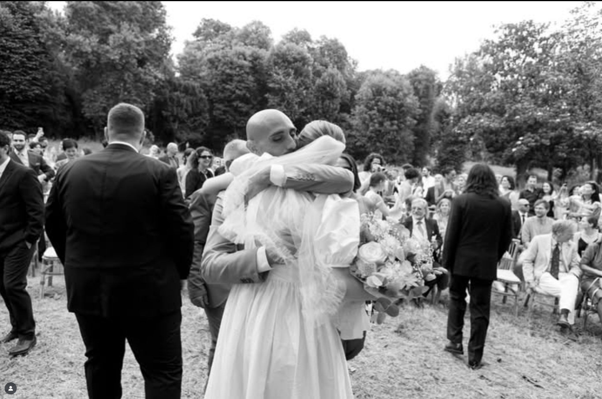 Una coppia di sposi si abbraccia durante un matrimonio all'aperto, circondata da amici e parenti seduti e in piedi tra gli alberi.