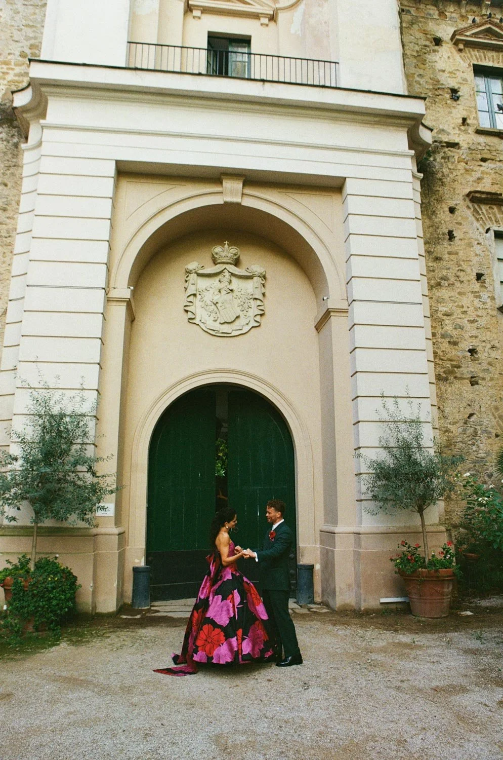 Una coppia di sposi si abbraccia davanti a un grande portone verde di un edificio storico, con decorazioni e stemma sopra l'arco, circondati da piante in vaso.