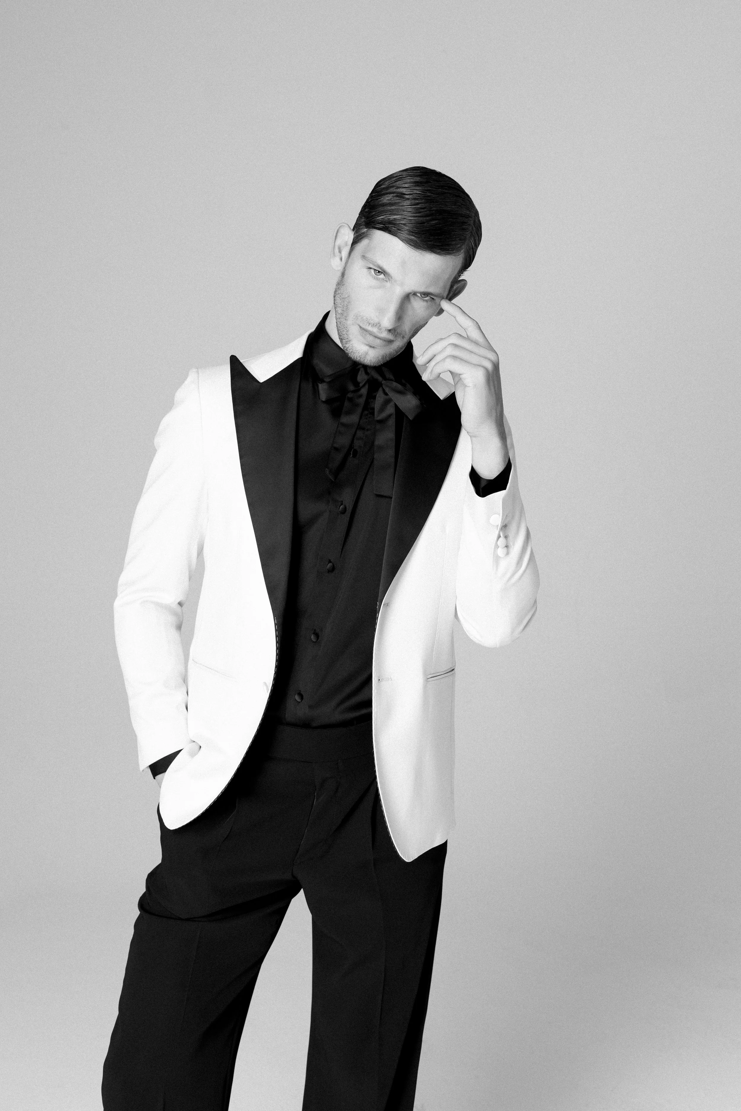 Ritratto di un uomo elegante con abito bianco e giacca nera, in posa pensierosa contro sfondo grigio, in bianco e nero.