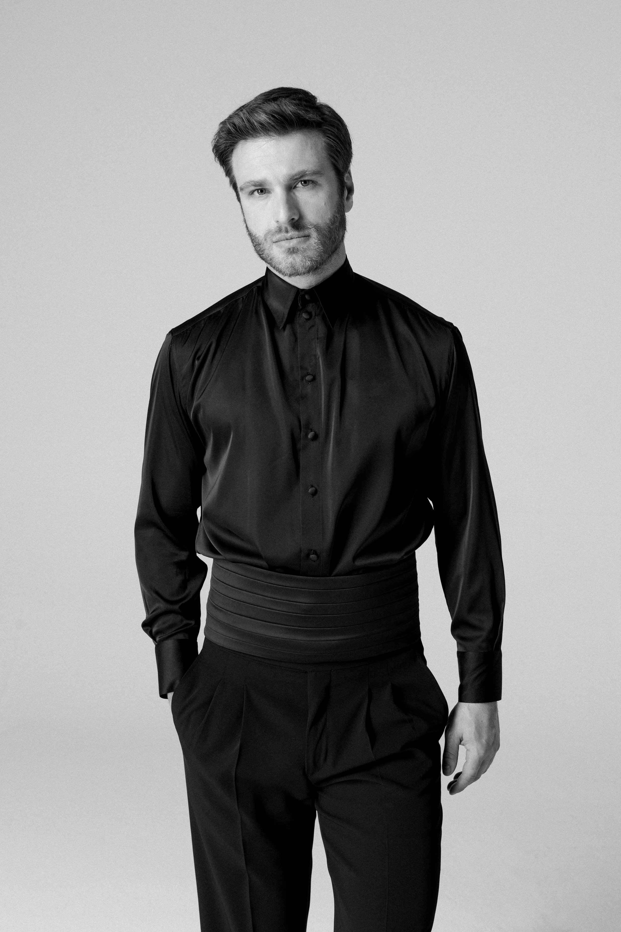 Uomo in abbigliamento nero, con camicia lucida e pantaloni, in posa contro sfondo neutro.