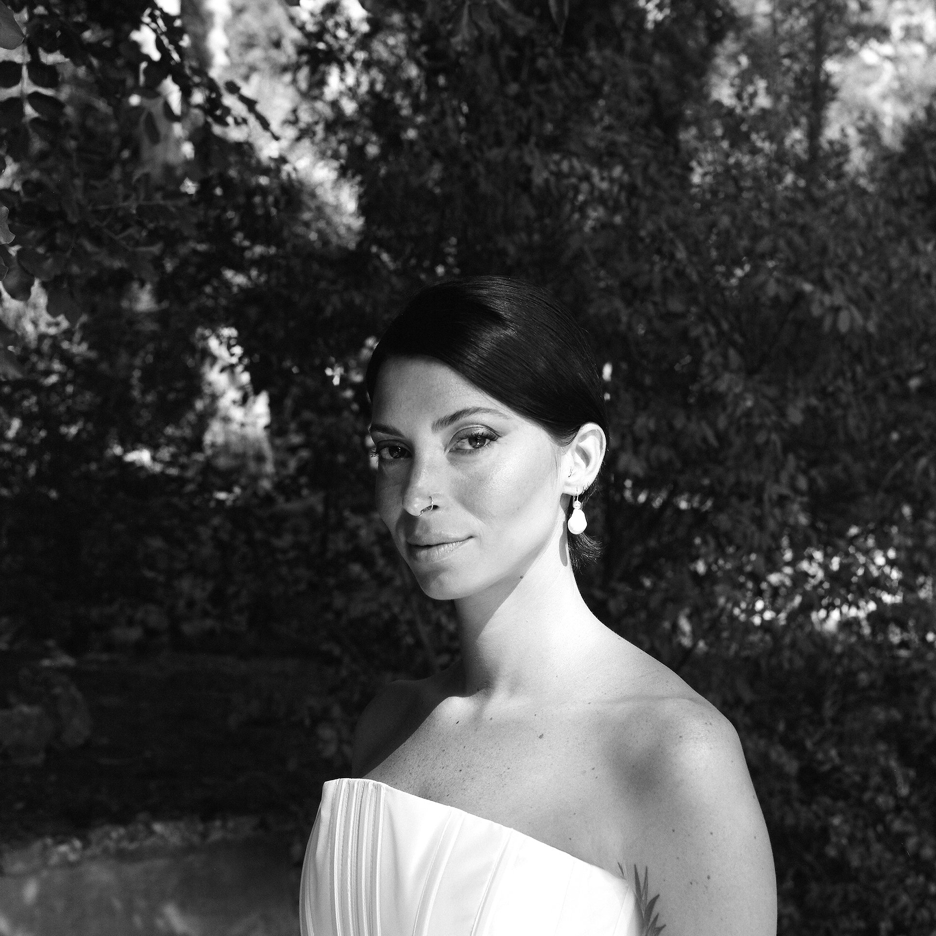 Ragazza con capelli neri corti, acconciatura liscia, indossa orecchini pendenti a goccia e un vestito scollato bianco, sullo sfondo un albero folto in un ambiente naturale.