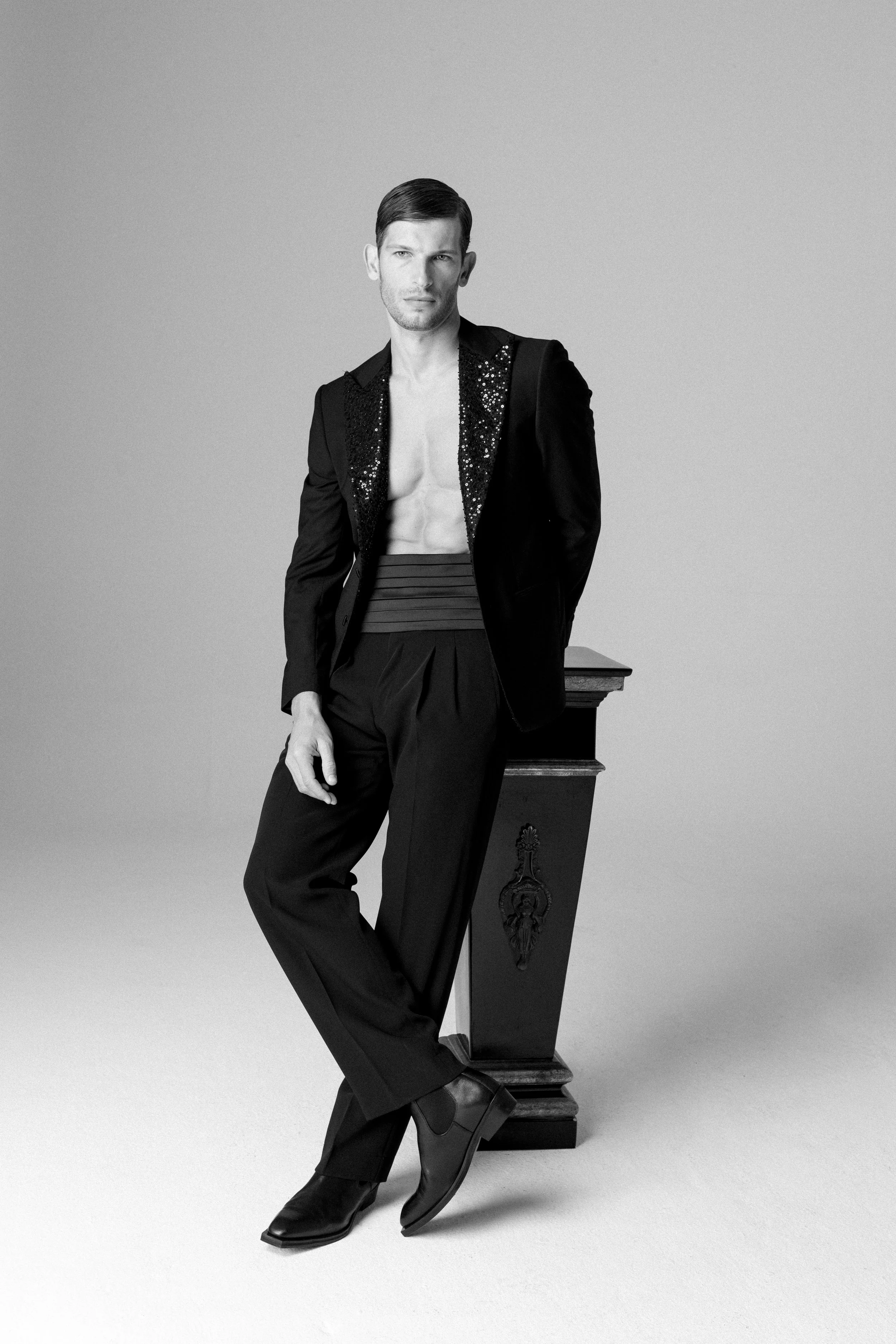 Uomo che indossa un completo nero con giacca decorata e pantaloni larghi, senza maglia, posizionato con una mano sul fianco e appoggiato a una scrivania in legno, in uno studio fotografico.