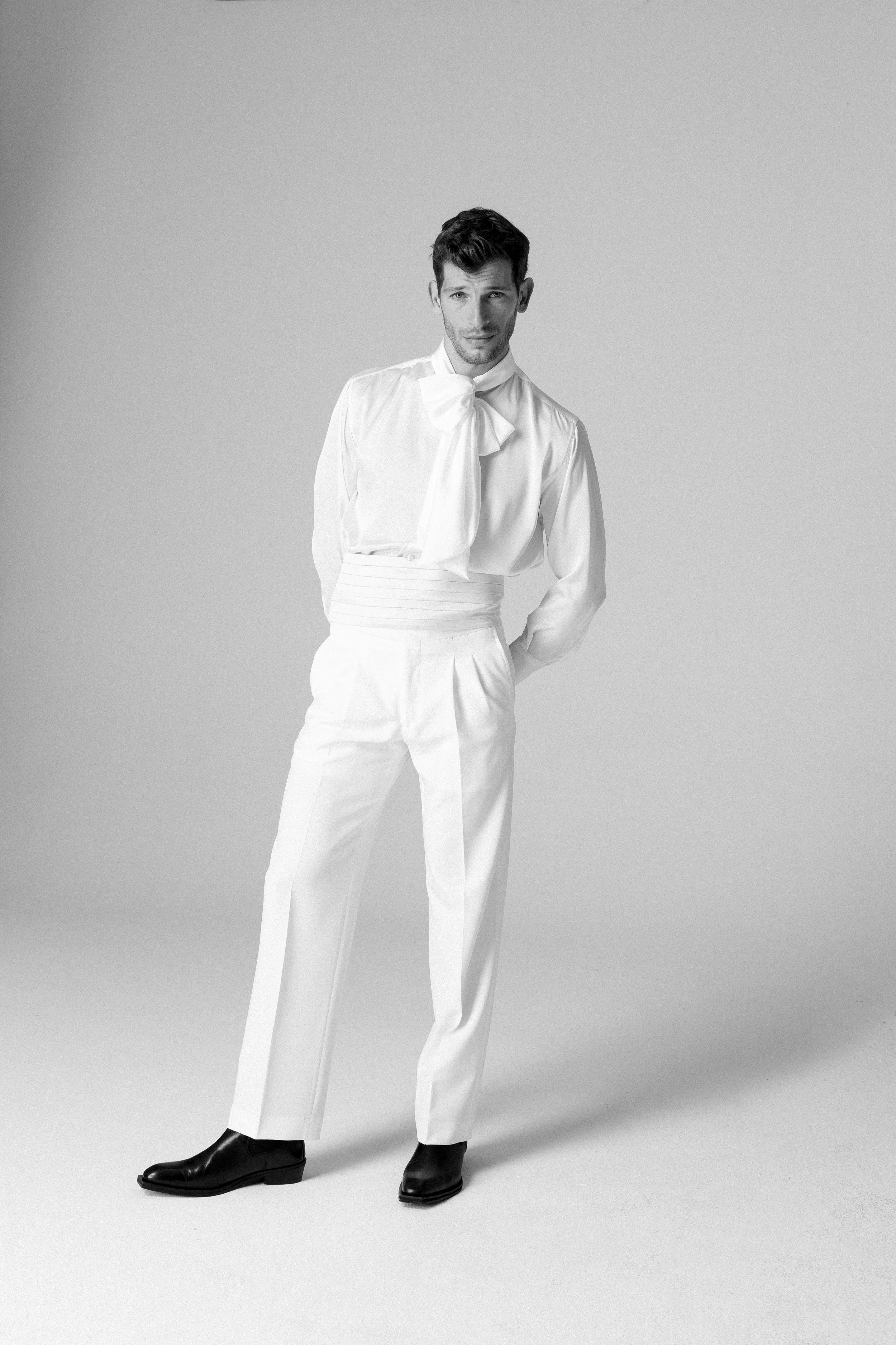 Uomo in abiti bianchi, pantaloni e camicia con cravatta, posa in piedi su sfondo neutro, vestito elegante e stile classico.