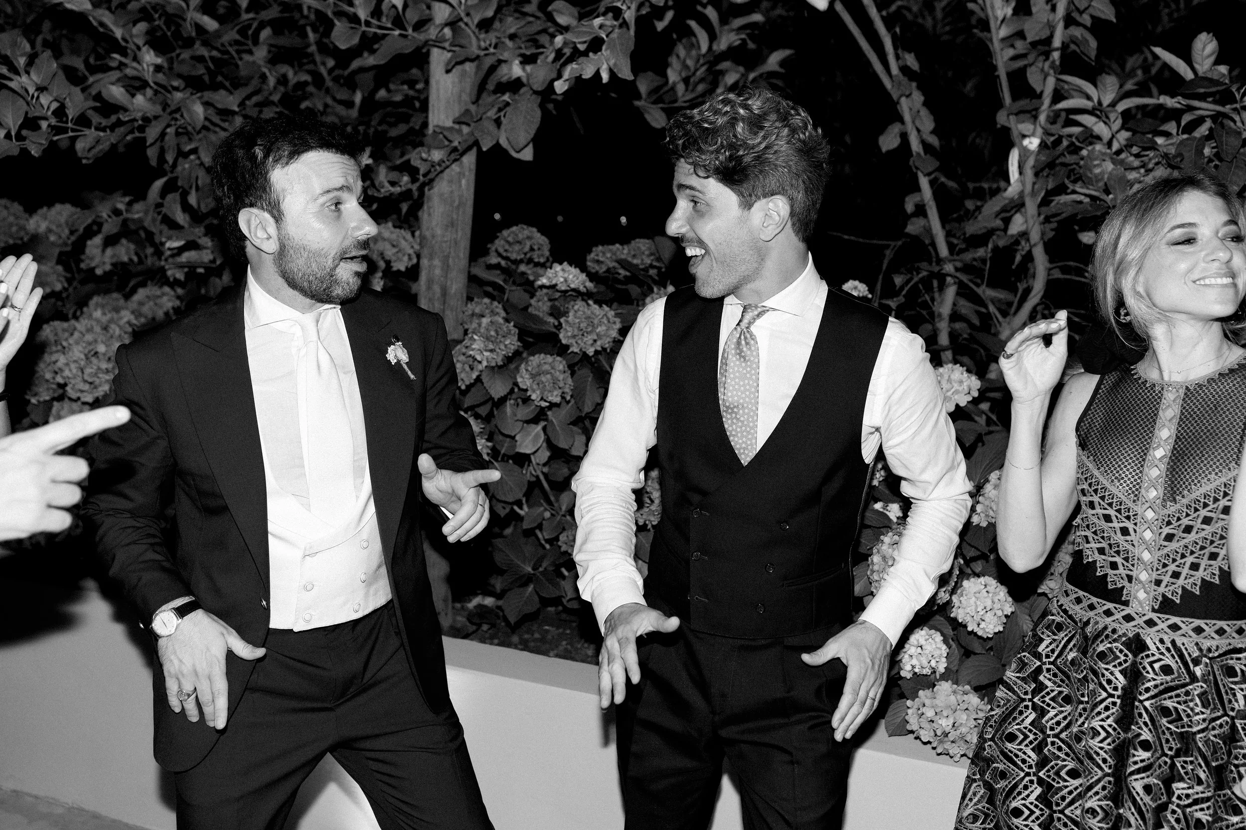 Tre persone che bailano e ridono durante una festa, con uno sfondo di fiori e foglie