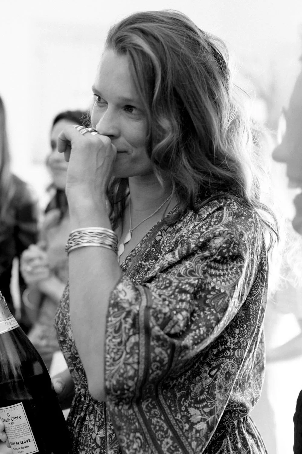 Donna sorridente con capelli mossi e accessori, che indossa un abito con motivo decorato, in un contesto di gruppo, con una mano vicino alla bocca.