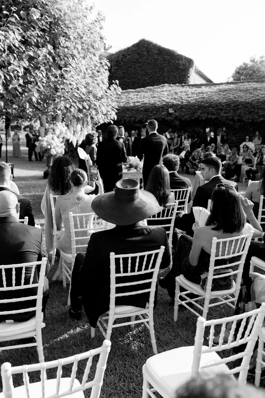 Cerimonia di matrimonio all'aperto con ospiti seduti su sedie bianche, coppia di sposi al centro, al tramonto o in prime luci, con una grande pianta e edifici sullo sfondo.