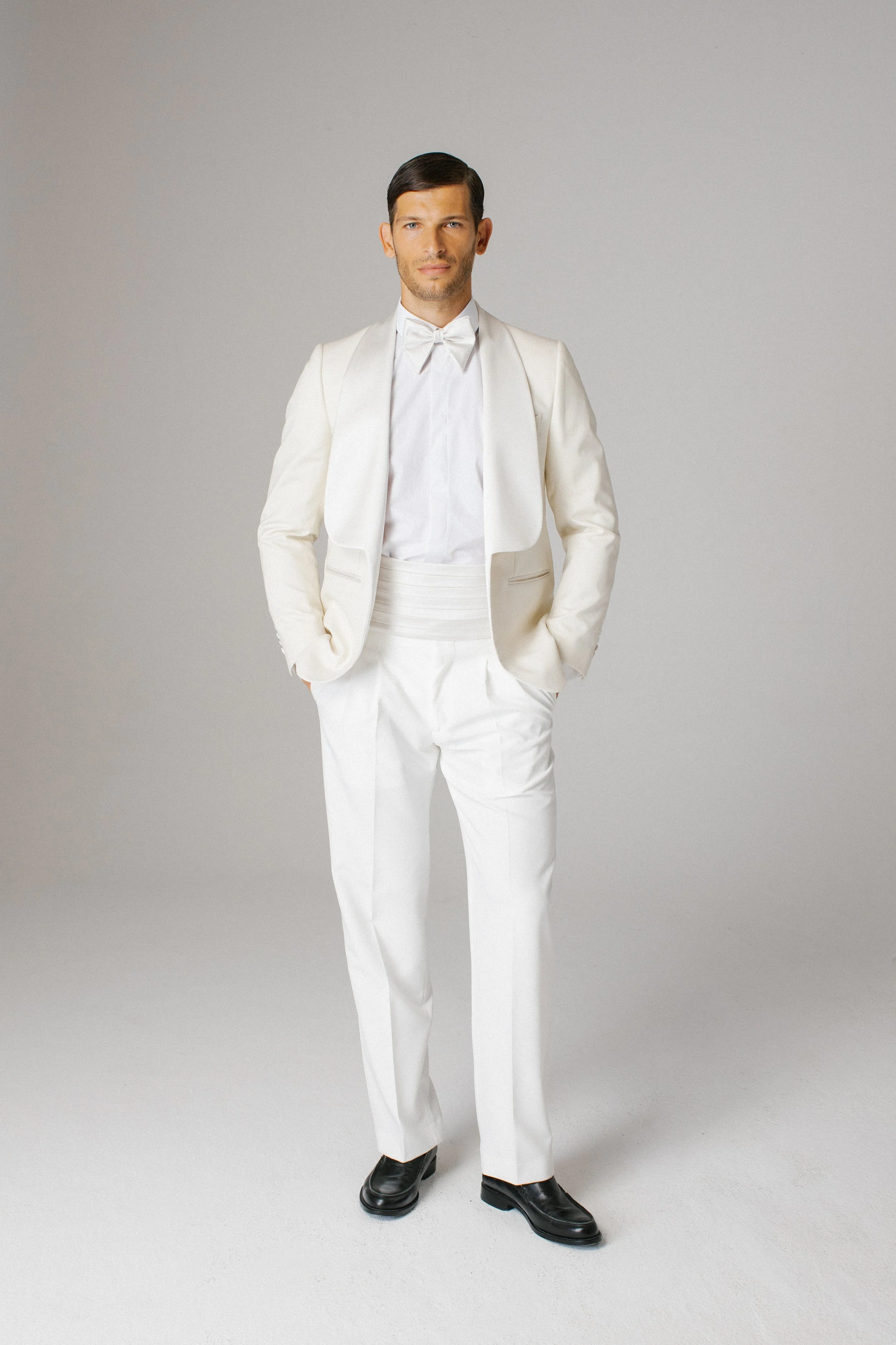 Uomo in elegante completo bianco con cravatta, posa con mani in tasca in uno studio con sfondo neutro.