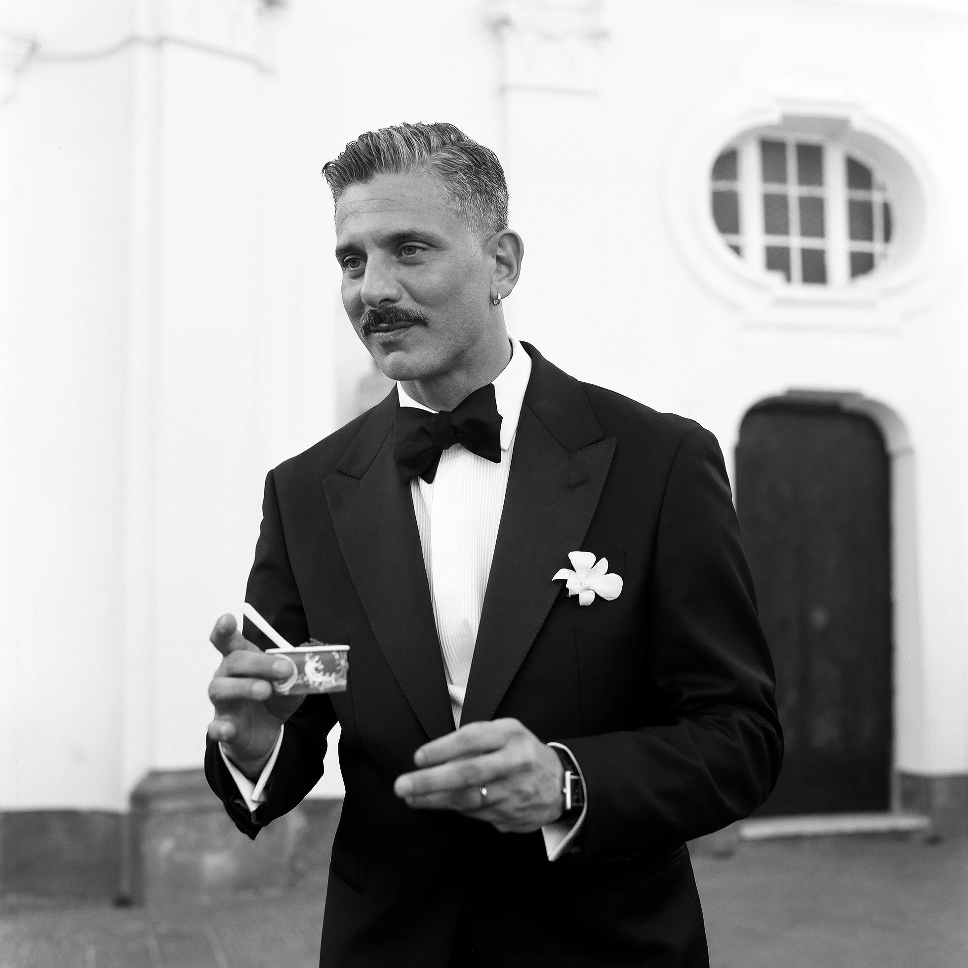 Uomo in abito da smoking con fiore sulla giacca, con baffi e orecchino, in una stanza. 