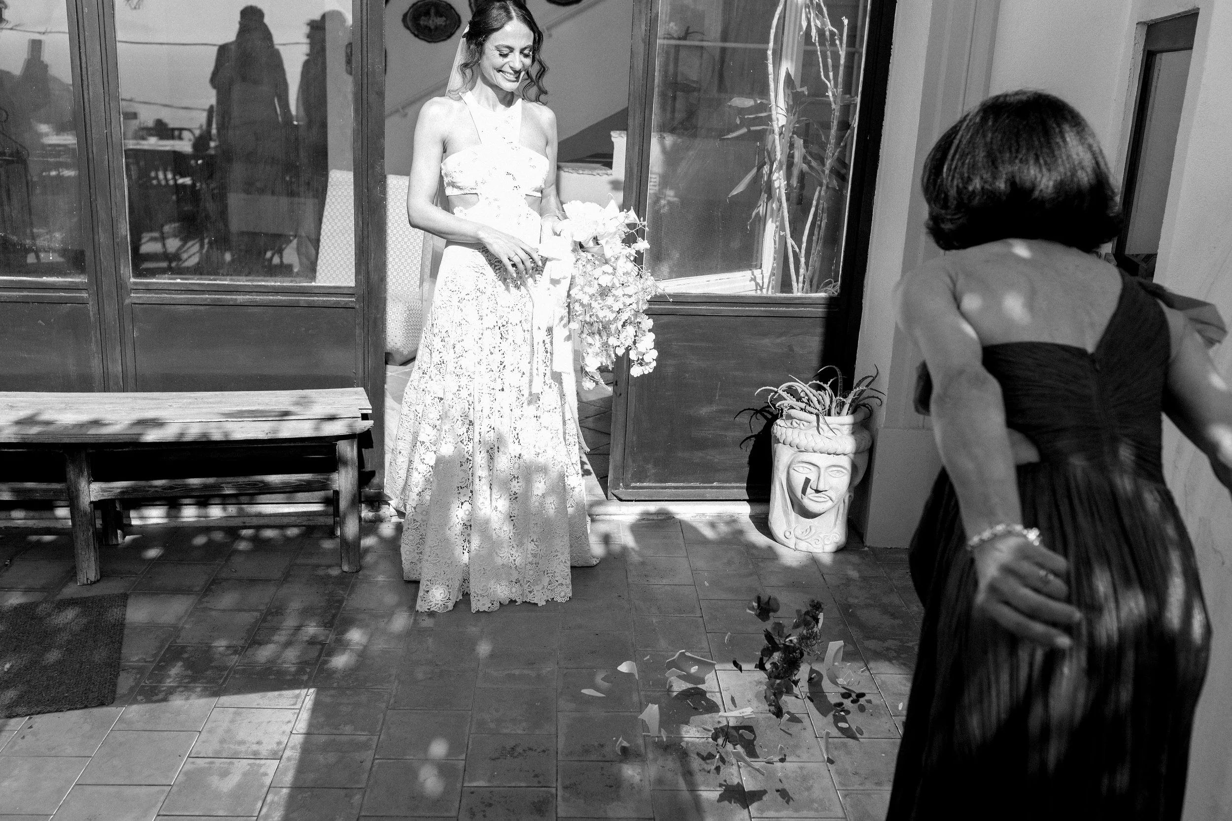 Una giovane donna in abito da sposa sorride mentre entra in una stanza, mentre un'altra donna, con il volto nascosto, si sta voltando verso di lei, in un'atmosfera di festa.