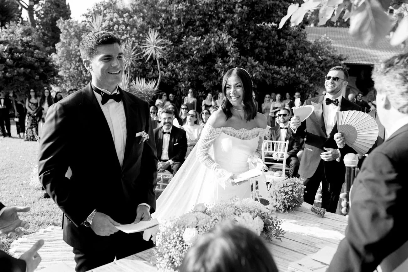 Gli sposi durante il matrimonio, con amici e testimoni seduti all'aperto, con alberi e un edificio sullo sfondo.