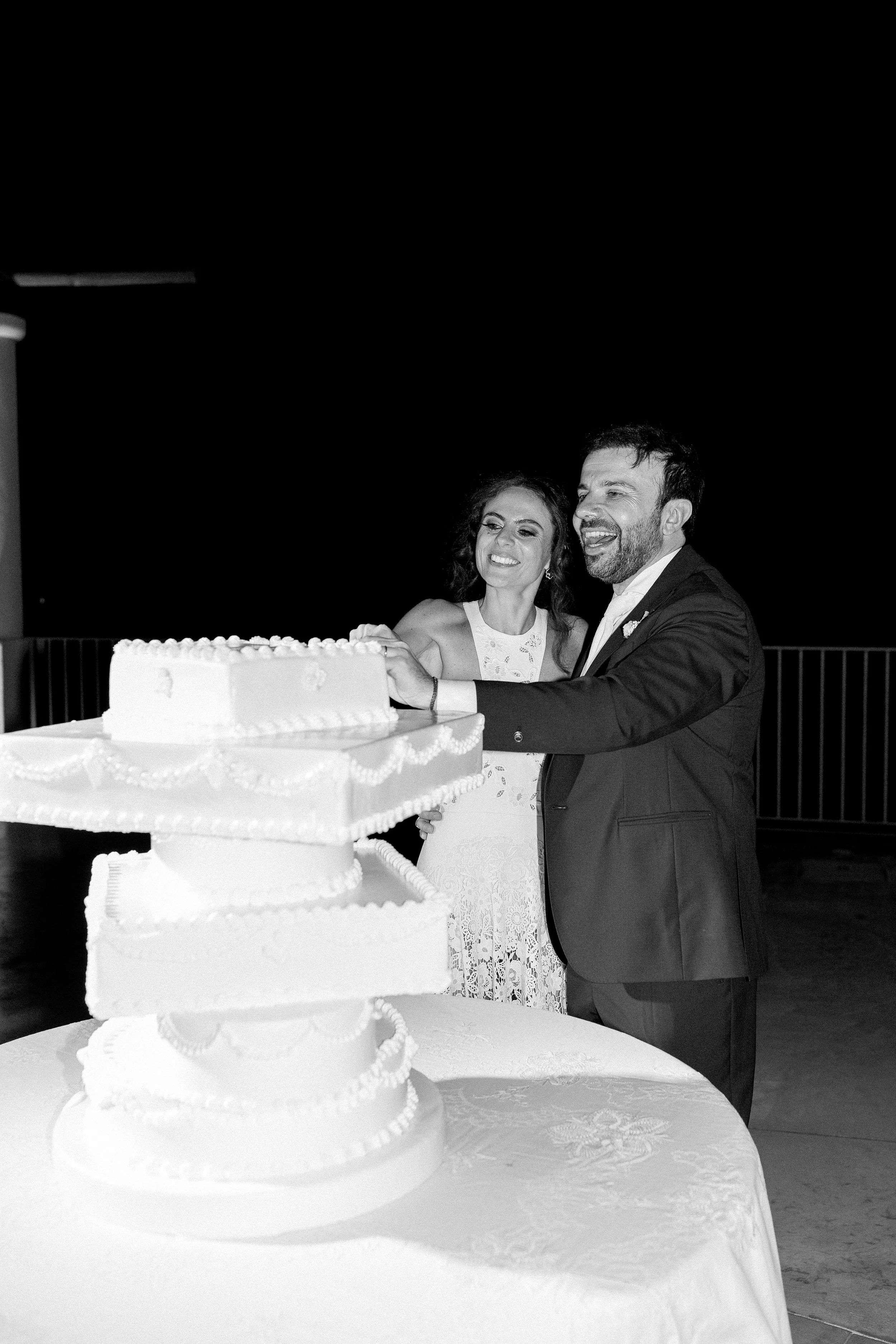 Una coppia di sposi in abito formale taglia una torta di nozze bianca decorata con piccole perline e dettagli eleganti, durante una festa di matrimonio notturna.