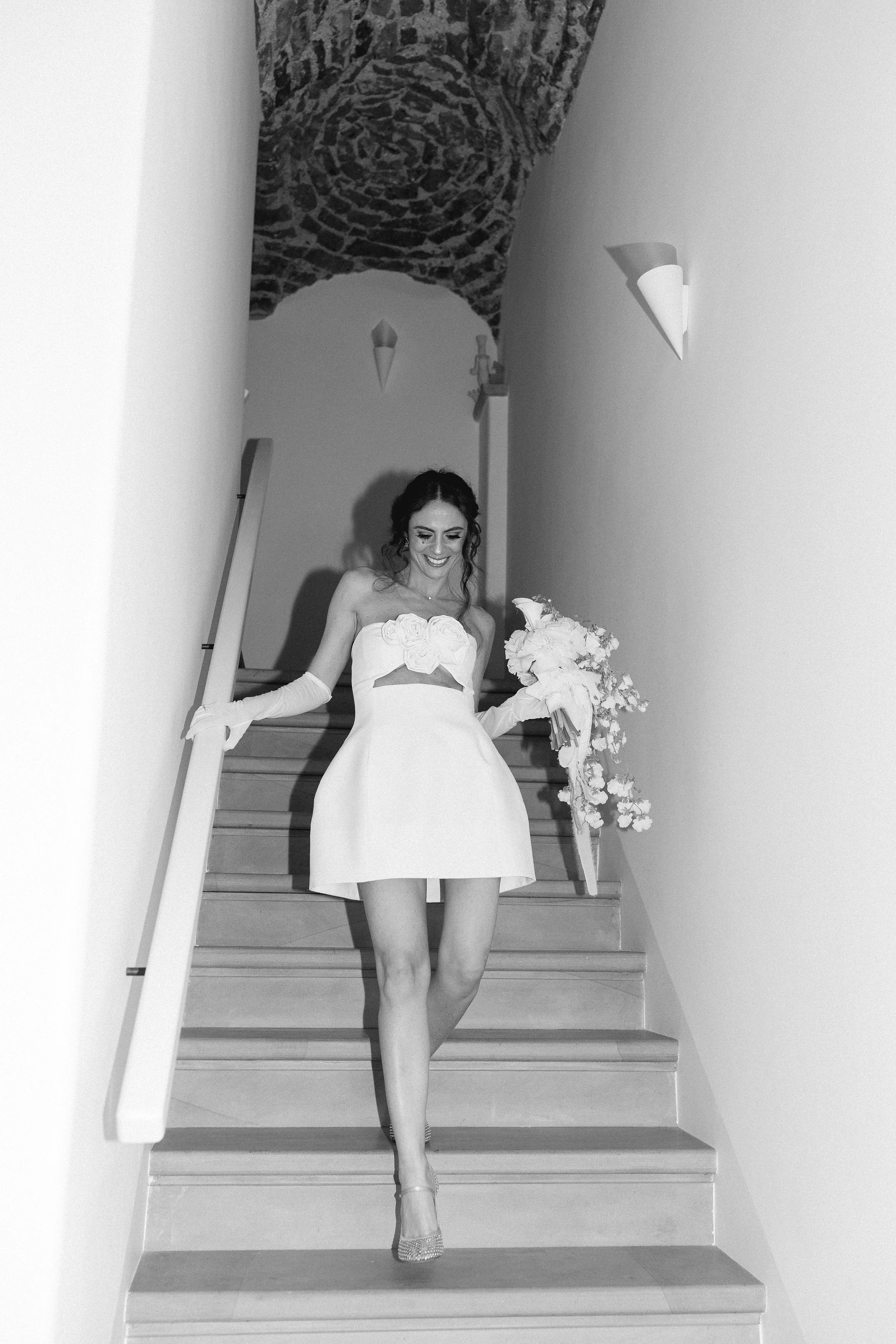 Una donna vestita in abito bianco con un mazzo di fiori in mano, scende le scale all'interno di un edificio.